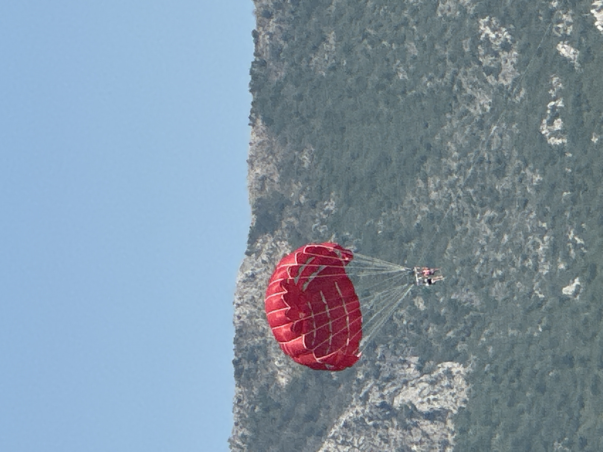 A parasailer with a red canopy is airborne against a backdrop of a rocky, tree-covered mountainside under a clear blue sky. Two individuals are visible in the harness suspended beneath the canopy. The image appears to be taken from a distance, possibly from a boat or the shore, looking up at the parasailer. The location is identified as Boreti, Montenegro.