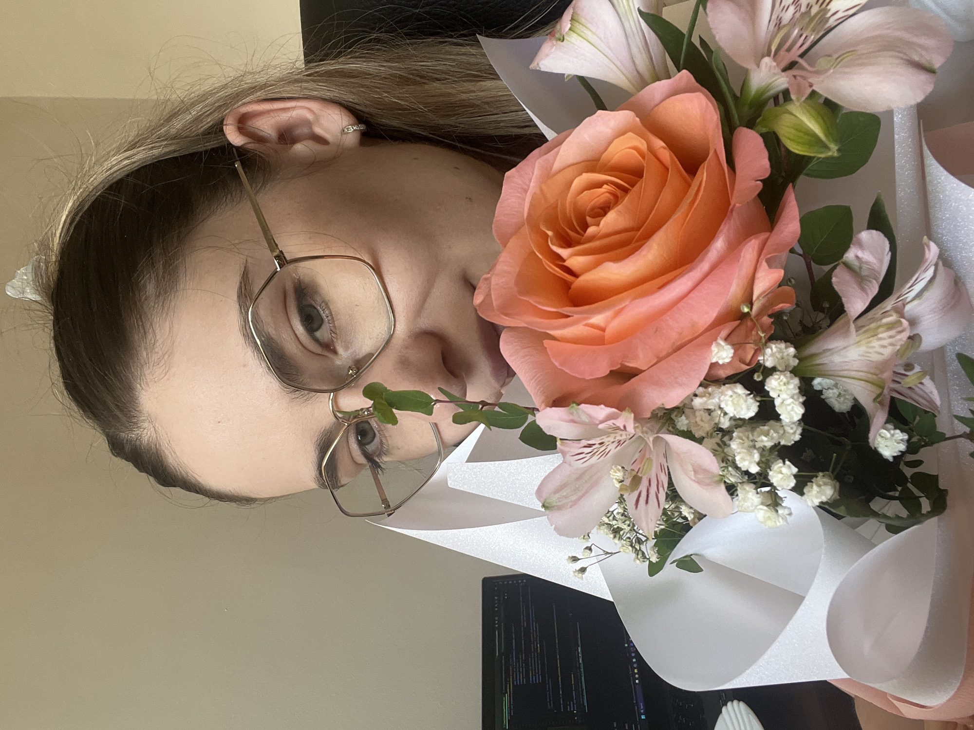 An eye-level, medium shot captures a young Caucasian woman holding a bouquet of flowers in front of her face. She is wearing large, gold-rimmed, geometric-shaped glasses and has light brown hair pulled back. A single small earring is visible on her right ear. The bouquet is composed of a large, peach-colored rose, several pale pink alstroemeria, and small white flowers, wrapped in white paper. To the left of the woman, a laptop screen displays lines of code. The background is a plain, light-colored wall and a black chair. The lighting is natural and soft, suggesting an indoor setting. The focus is on the woman's eyes and the flowers.