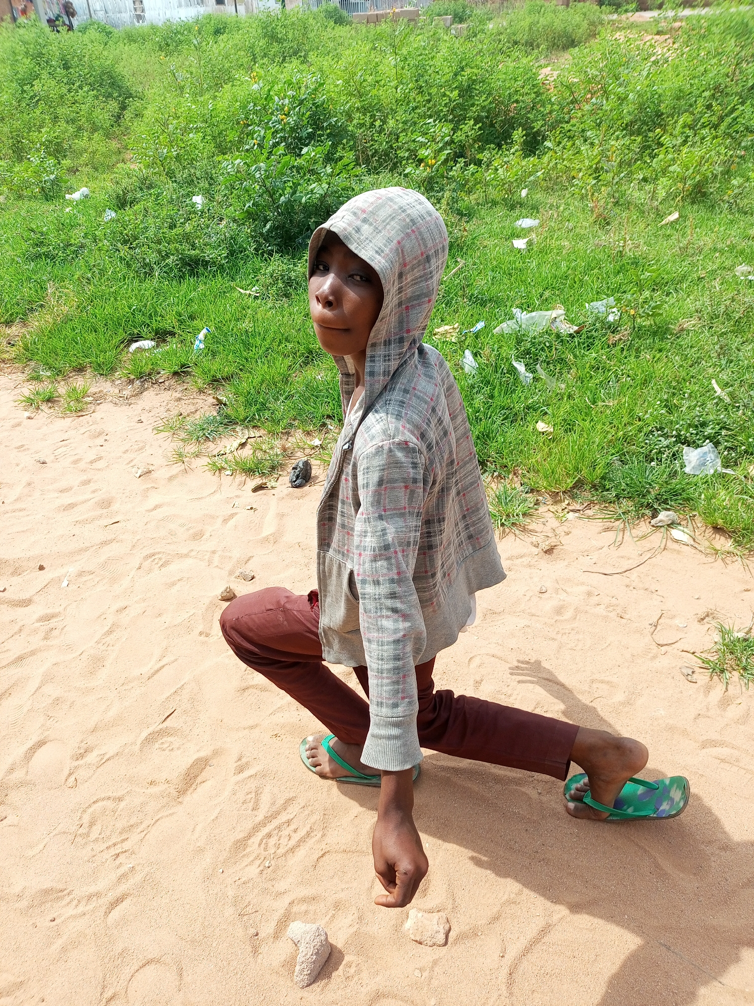 A young person is kneeling in the sand, looking towards the camera. They are wearing a checkered hooded jacket and brown pants, with green patterned flip-flops on their feet. The background shows a grassy area with some scattered litter. The scene appears to be outdoors in a dusty, possibly rural or suburban environment within Tempure, Nigeria. The lighting suggests it is daytime.