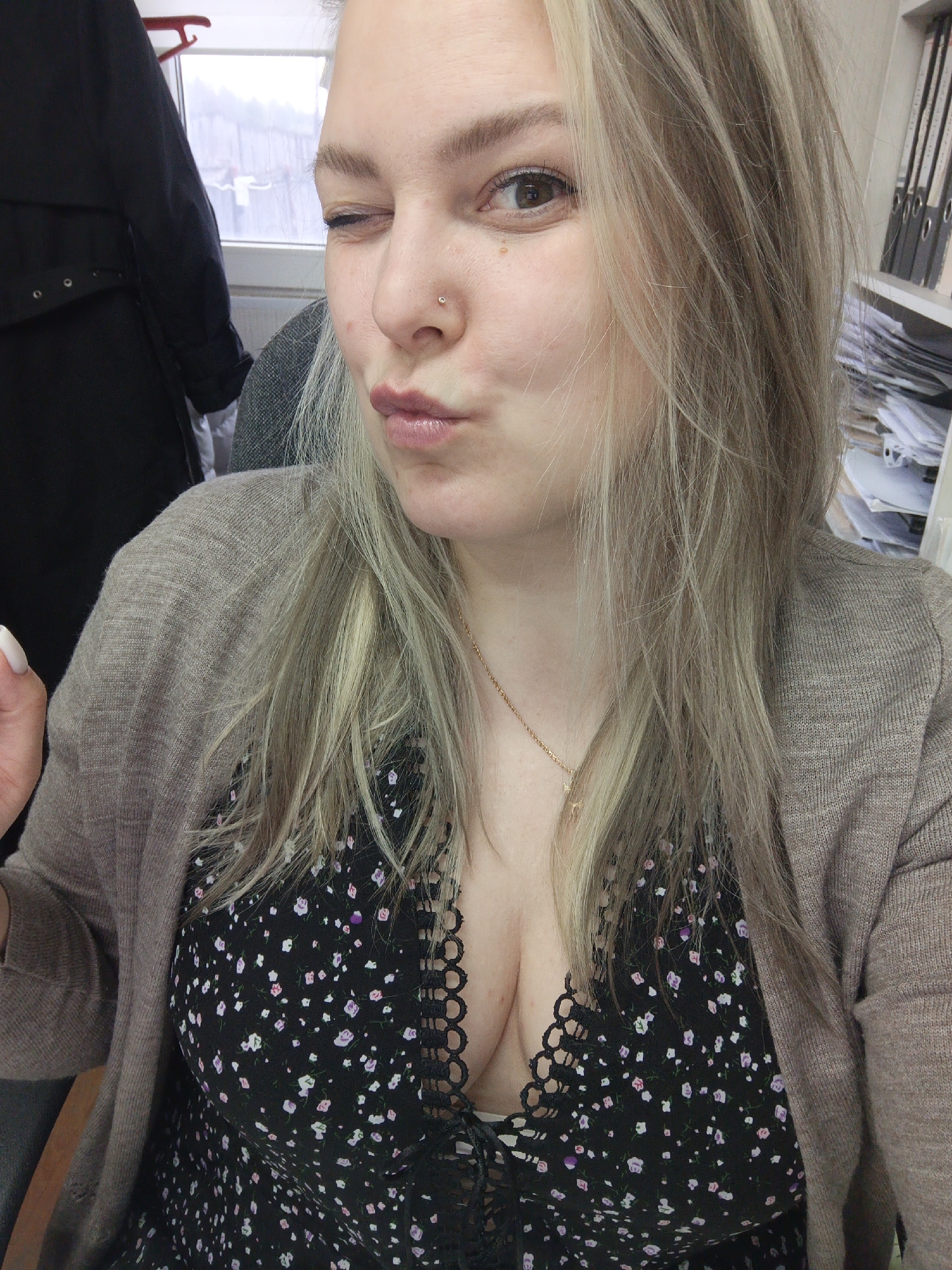 A woman with blonde hair and a nose piercing is winking at the camera and pursing her lips. She is wearing a black dress with a floral pattern and a tan cardigan. A gold necklace is visible around her neck. In the background, a black coat hangs on a rack near a window. To the right, there is a cluttered desk with stacks of papers and binders. The exact location in the city of Noviy, Russia is not discernable from the image.