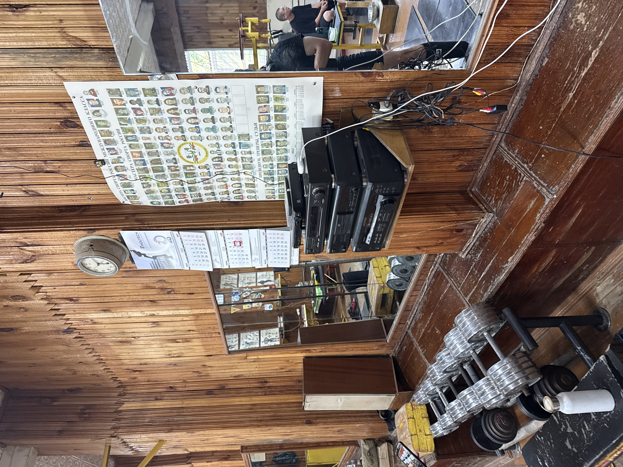 This image captures a scene within a gym, likely in Kazan, Russia, characterized by its rustic wooden paneling. In the foreground, a rack of chrome dumbbells is visible, along with several larger, black-plated weight discs. A white water bottle rests on a nearby black surface. To the left, a tall, light-colored cabinet with a dark wooden front panel stands against the wall.
Dominating the background on the wooden wall is a large poster featuring numerous portraits of individuals, likely athletes or esteemed figures, arranged in rows. Below this, a framed clock with Roman numerals hangs on the wall. Adjacent to the poster are calendars for the years 2025 and 2026. A large mirror on the right wall reflects the gym's interior, showing workout equipment and a person in the background. A stack of audio-visual equipment, including what appears to be multiple CD players or receivers, is mounted on a wooden shelf attached to the wall. Various wires and cables are connected to this equipment and hang loosely. The overall impression is of a well-used, older gym facility.