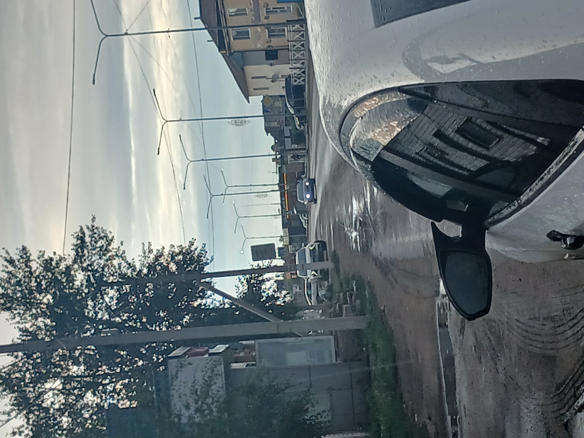 This is a street-level, medium shot image taken from inside a vehicle, looking outwards towards a wet road and buildings in Makinsk, Kazakhstan. The foreground is dominated by the side of a white vehicle, with its side mirror and part of a window visible. The window reflects a building with brickwork and multiple windows.
The road surface is visibly wet and appears to be unpaved or poorly maintained, with tire tracks and puddles. A sidewalk or verge runs alongside the road, with patches of grass and some debris.
On the opposite side of the road, there is a row of buildings. The closest building appears to be a single-story structure, possibly a shop or residential building, with a yellow facade and dark trim. Further down the street, more buildings are visible, including a multi-story structure that appears to be commercial. There are streetlights lining the road, and overhead power lines are also present.
Several vehicles are on the road. A dark-colored car is visible in the distance, approaching the viewer. Another white SUV is parked on the side of the road. There are also other cars further down the street, but they are less distinct. Trees with dark foliage are present on the left side of the image, partially obscuring a building that has a gray, textured facade and a large window. The sky is overcast with a light gray tone.