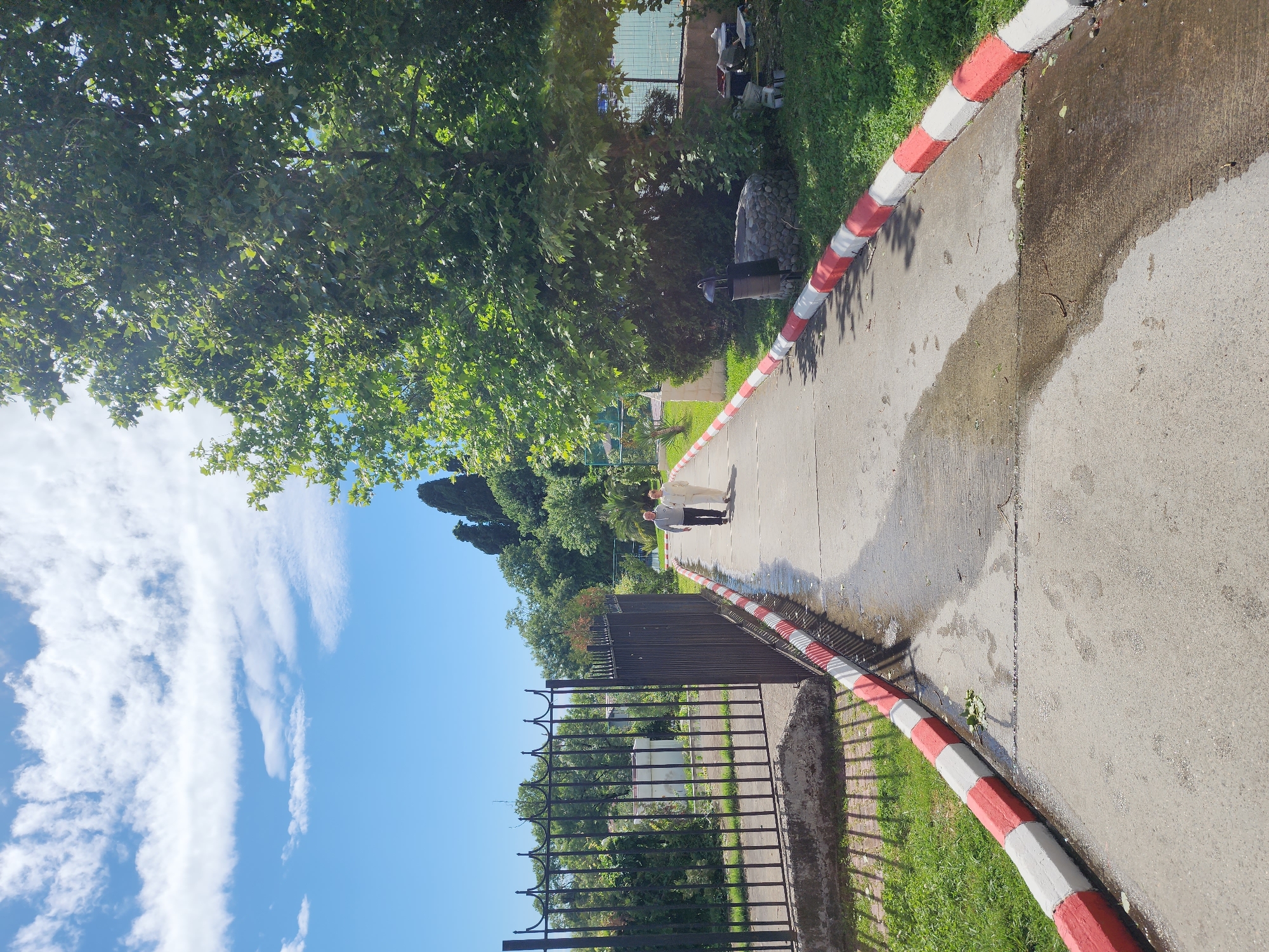The image depicts a street scene in Sochi, Russia, showcasing a paved walkway bordered by a distinctive red and white curb. The walkway appears to be slightly wet, suggesting recent precipitation or cleaning. Two individuals are visible further down the path, seemingly walking together. A black iron fence runs alongside the walkway on one side, separating it from a grassy area. Lush green trees and blue sky can be seen overhead.