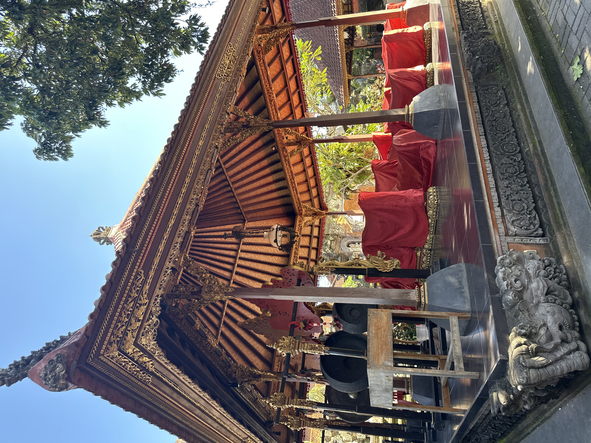 An outdoor, low-angle shot captures an ornate, open-air pavilion with a thatched roof in Ubud, Indonesia. The pavilion, likely a part of a temple complex, features intricate golden detailing along its eaves and roof structure. Inside, a set of large gongs rests on a wooden stand on the left. The rest of the pavilion's interior is covered by vibrant red fabric draped over unseen objects, possibly seating or ritualistic items. The pavilion's platform is elevated and tiled in black. The base of the structure is adorned with detailed stone carvings, including a mythical creature resembling a dragon or lion. In the background, lush green trees and other traditional Balinese buildings with tiled roofs are visible under a clear blue sky. The sunlight casts shadows, indicating it is daytime. No people are present in the scene.