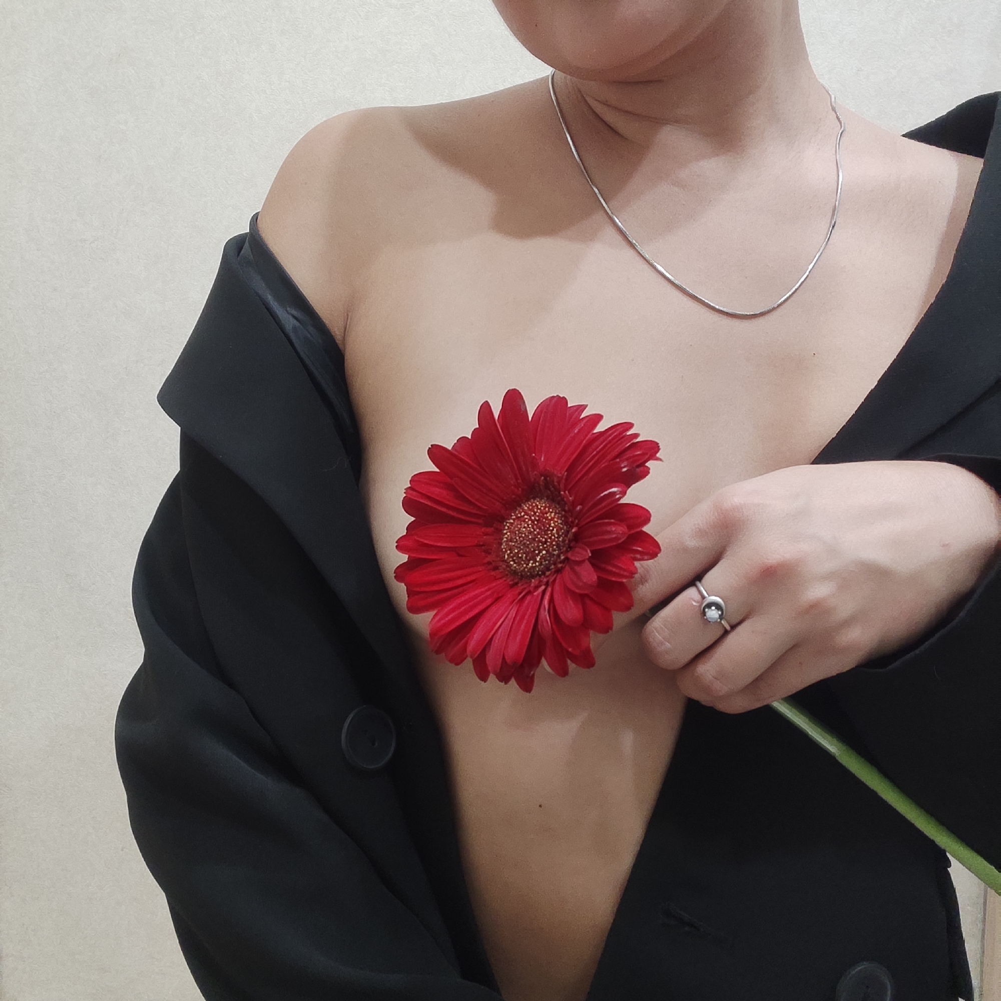 A person is photographed indoors. They are wearing a black jacket that is open, revealing their chest area. A large, red gerbera daisy covers their chest. The person is also wearing a silver necklace and a ring on their finger. They are holding the stem of the flower. The background is a plain wall. There is no indication of the city Yekaterinburg, Russia in the image.