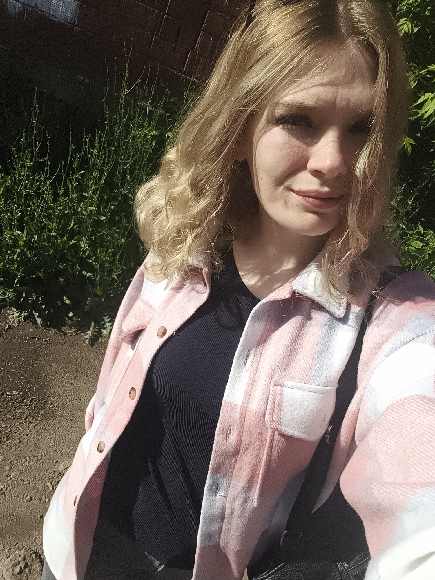 The image features a young woman taking a selfie outdoors in Izhevsk, Russia. She has blonde, wavy hair and a fair complexion, looking directly at the camera with a subtle, pleasant smirk. She is wearing a pink, white, and light blue plaid shacket or jacket over a black ribbed top. The jacket has visible light brown buttons and a single chest pocket. A black bag strap is visible over her right shoulder.
The background consists of green foliage and tall weeds, suggesting a park or garden setting. A dark brick wall is partially visible in the upper left portion of the frame. The ground in the foreground and mid-ground is brown and earthy. The bright sunlight indicates a clear, sunny day, casting some shadows across her face and the environment. There is no visible text in the media file.