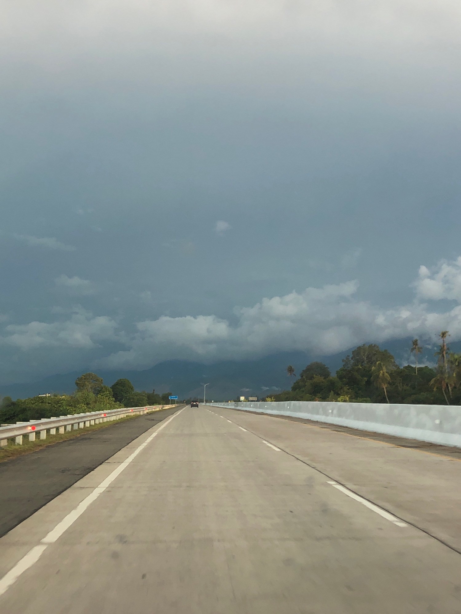 The image depicts a highway in the city of Simomlhok, Indonesia, under a cloudy sky. The highway is paved with concrete and has white lane markings. On the left side of the highway, there is a metal guardrail with red reflectors. Beyond the guardrail, there are trees and vegetation. On the right side of the highway, there is a concrete barrier. In the distance, there are mountains. A black car is visible on the highway in the distance. The sky is filled with dark, heavy clouds, suggesting an impending storm.
