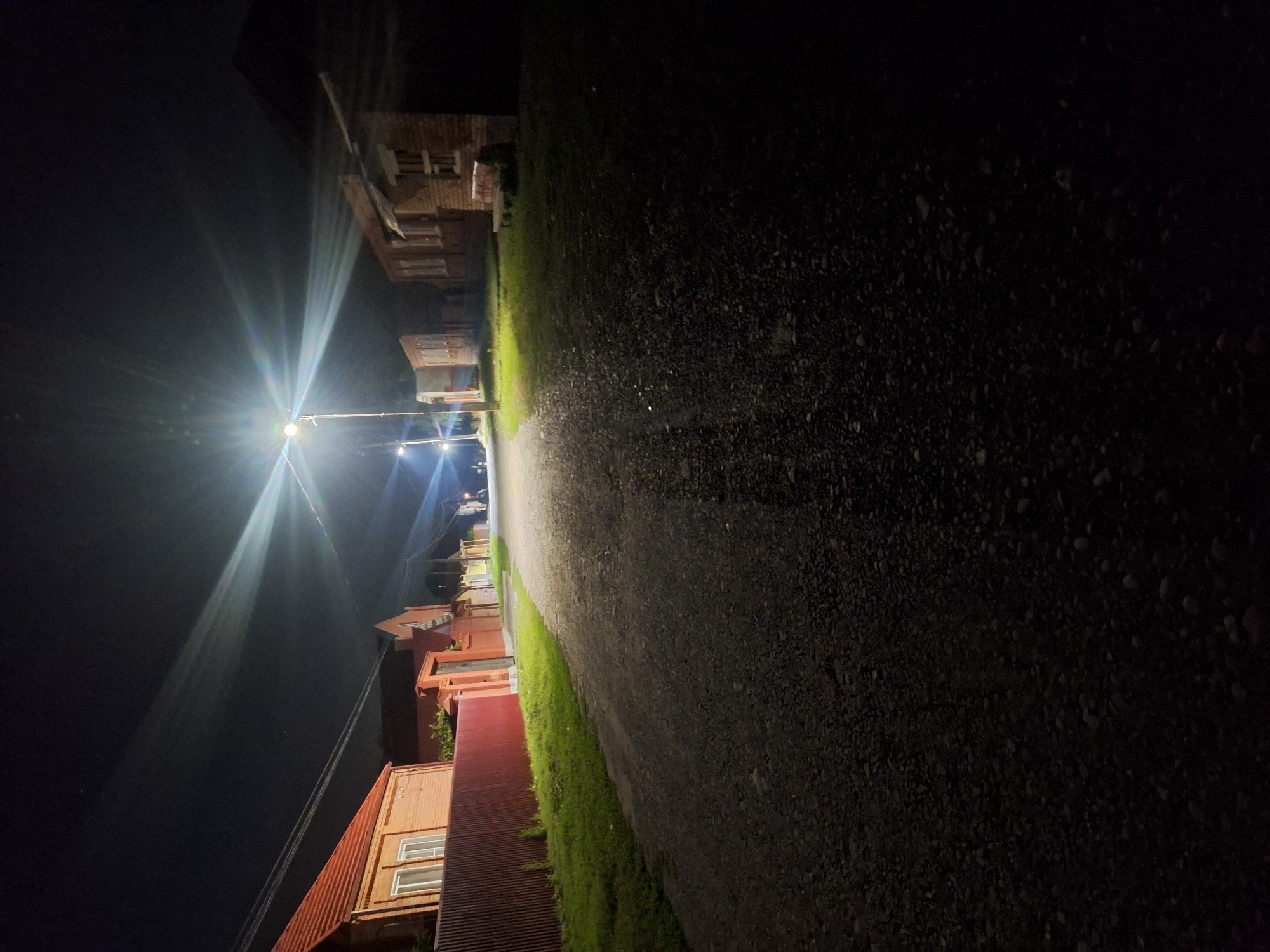 This is a nighttime photograph of a street in Chermen, Russia, illuminated by a bright streetlamp. The street is unpaved and appears to be made of gravel. On the left side of the street, there are brick buildings with red tiled roofs and a red wooden fence. On the right side of the street, there are also brick buildings, some of which appear to be under construction or dilapidated. The sky is dark, indicating it is nighttime. The streetlamp casts long, starburst-like rays of light across the scene. There are no people or vehicles visible in the image. The overall impression is one of a quiet, rural street at night.