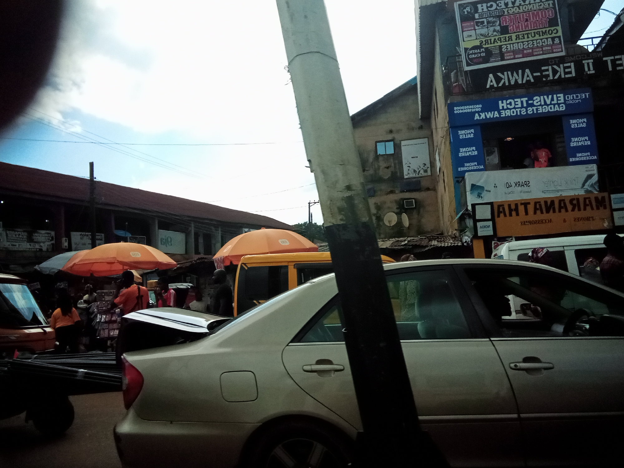 The image captures a street scene in Nibo, Nigeria, under an overcast sky. In the foreground, a silver sedan is partially visible from the side, its rear end slightly obscured by a dark, vertical pole. To the left of the sedan, the rear of a dark truck or trailer bed is visible, loaded with metal rods. In the background, a bustling market environment unfolds. Several orange and grey umbrellas provide shade over stalls. People, some wearing bright orange and red shirts, are seen interacting around these stalls, suggesting commercial activity. Buildings line the street, with some featuring prominent signage. One building displays a large blue sign with white lettering, likely indicating a business, and above it, several posters or advertisements are affixed to the wall. The overall impression is of a lively, everyday scene in a Nigerian city.