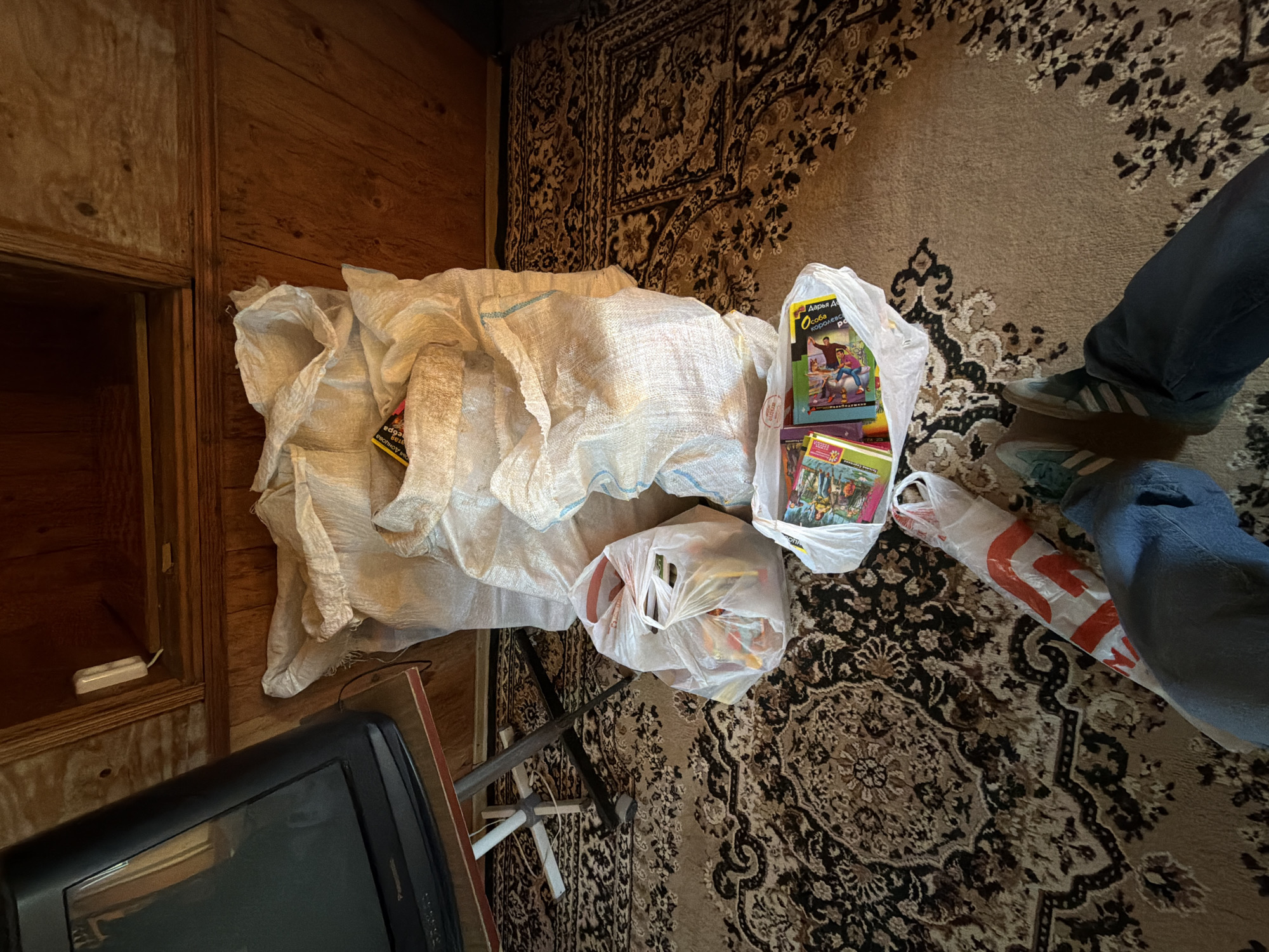 The image shows a cluttered indoor scene, likely in a residential setting. The floor is covered with a patterned rug. Several objects are scattered around, including a large sack-like object, two plastic bags filled with items, and a television set in the lower left corner. A pair of legs wearing jeans and Adidas shoes is visible in the upper right corner, indicating a person standing nearby. The scene takes place in Kamyshin, Russia.