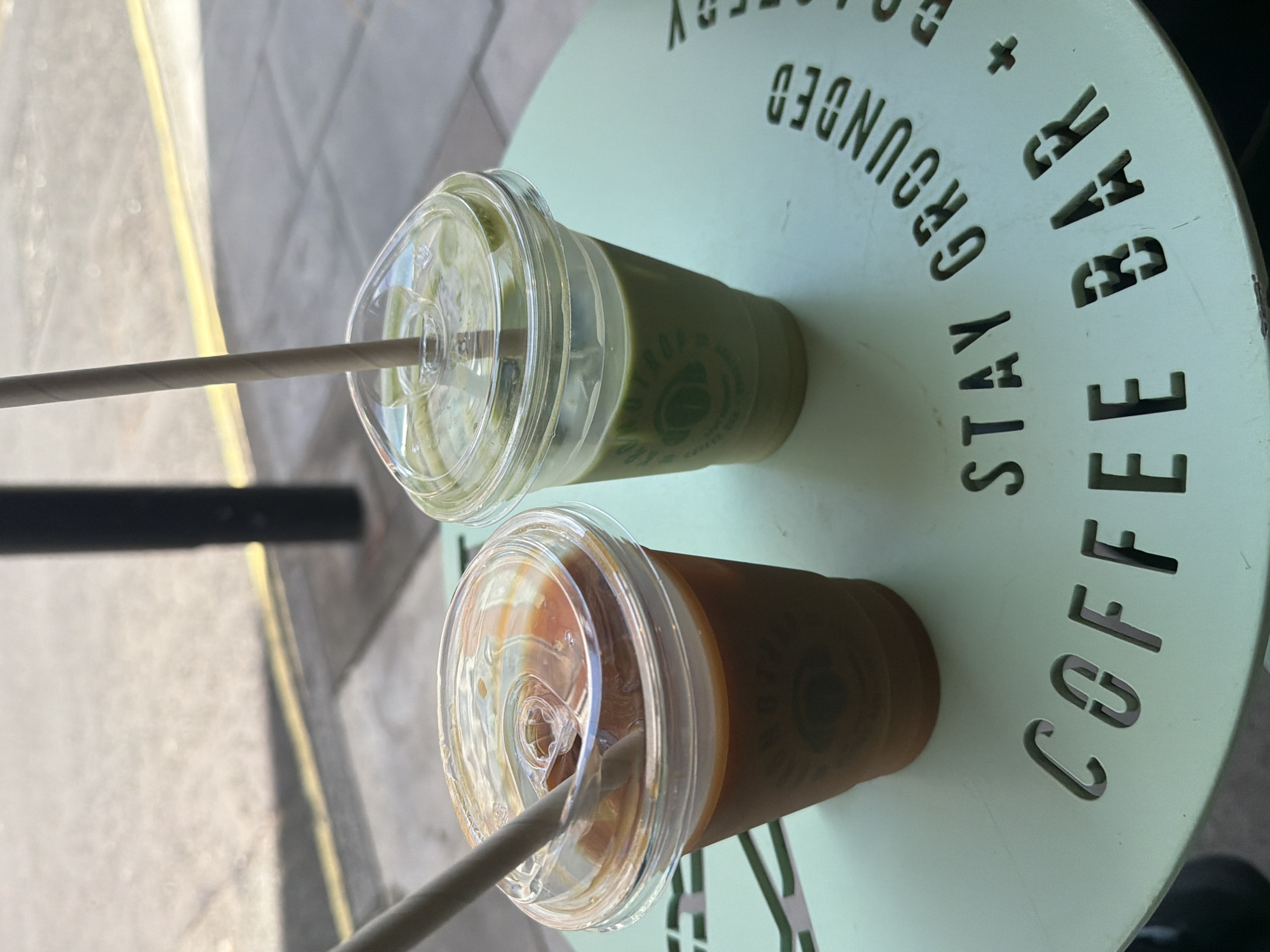 The image shows two takeaway drinks on a pale green table with the words "STAY GROUNDED + COFFEE BAR" cut out of the table surface. One drink is a green-colored beverage, possibly matcha, and the other is brown, likely coffee. Both have clear plastic lids with paper straws inserted. The location is likely London, United Kingdom, based on the style of coffee establishment and general urban aesthetic. The scene is presented from an overhead perspective. The table is situated outside, evidenced by the pavement visible in the background.
