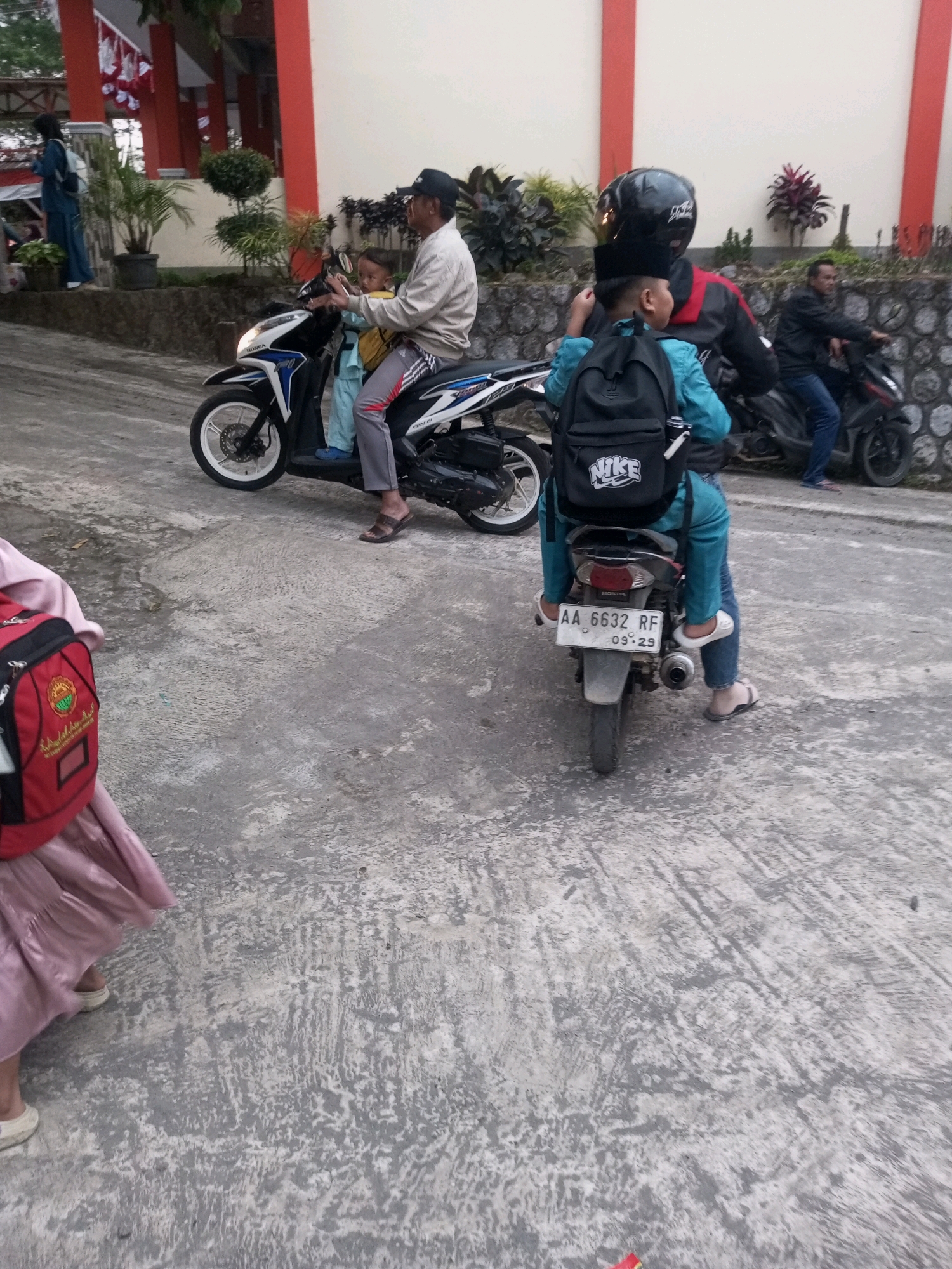 The image depicts a street scene in Krasak, Indonesia. In the foreground, on the left, a person wearing a pink dress and a red backpack is partially visible, walking away from the camera. In the middle ground, a man wearing a cap and light-colored shirt is riding a motorcycle, with a child in a light blue outfit sitting in front of him. The motorcycle is white and blue. Behind them, a person wearing a black helmet and a blue outfit with a black backpack is riding another motorcycle. This motorcycle has a license plate that reads "AA 6632 RF 09.29". In the background, another person is riding a motorcycle. The environment consists of a paved road and a stone wall behind it, with some greenery and buildings visible. The overall lighting suggests it is daytime.
