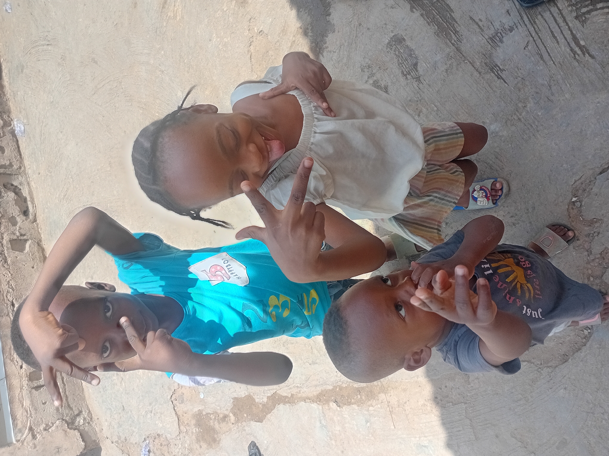 Three children are captured in a playful pose on an outdoor concrete surface, appearing sideways due to the image orientation. In the center, a young girl with braided hair wears a light-colored, ruffled top and striped bottoms, along with light blue sandals featuring a cartoon character (resembling SpongeBob). She grins widely, sticking out her tongue and making a peace sign with both hands.
To her left, a boy in a bright turquoise t-shirt and dark shorts looks towards the camera. He forms a "shaka" or "hang loose" sign with his left hand and a peace sign with his right, framing his face. Visible text on his shirt, partially inverted, reads "HAPPY BOY".
To the girl's right, a younger boy in a dark grey t-shirt and dark shorts also looks up at the camera. He covers part of his mouth with one hand while forming a "shaka" sign with the other. His shirt features text that is partially legible as "just shine like the sun is go...". He wears light-colored sandals.
The setting is an open, sunlit area with a rough, worn concrete ground. Strong shadows suggest direct sunlight, indicating it is daytime. The children display lively and engaged expressions, with the girl being particularly expressive.