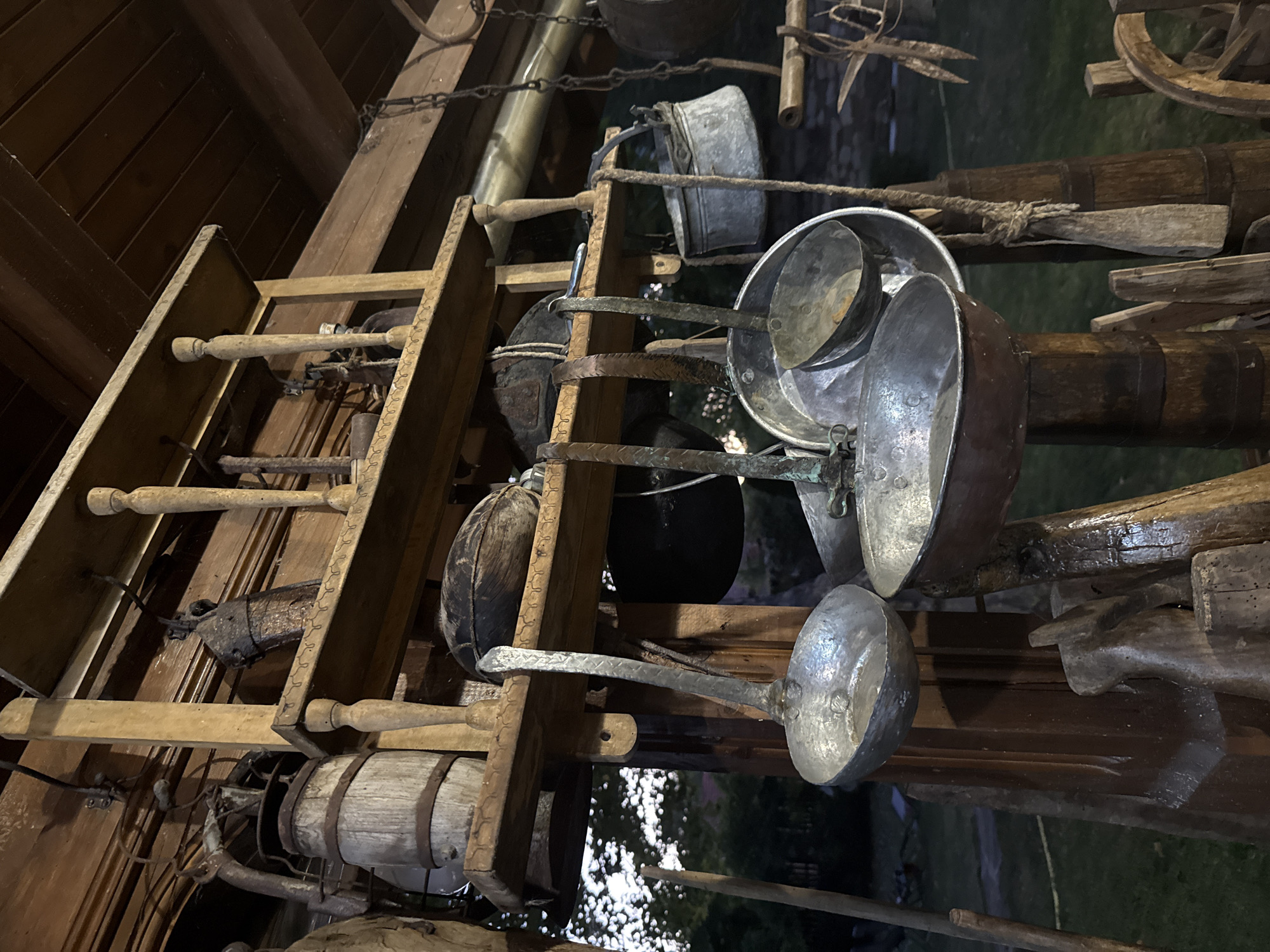 This image captures a display of antique kitchenware and tools, likely for a museum or historical exhibit, situated outdoors in Petrevene, Bulgaria. The collection is presented on wooden shelves and hanging from hooks and chains, suggesting an rustic and preserved atmosphere. The items include various metal ladles and pots, some appearing to be made of copper or tin. A wooden barrel is visible on the top shelf, alongside other rustic wooden objects. The background shows greenery, indicating an outdoor setting, possibly a yard or a public space. The lighting suggests it is either early morning or late evening. There are no people present in the image.