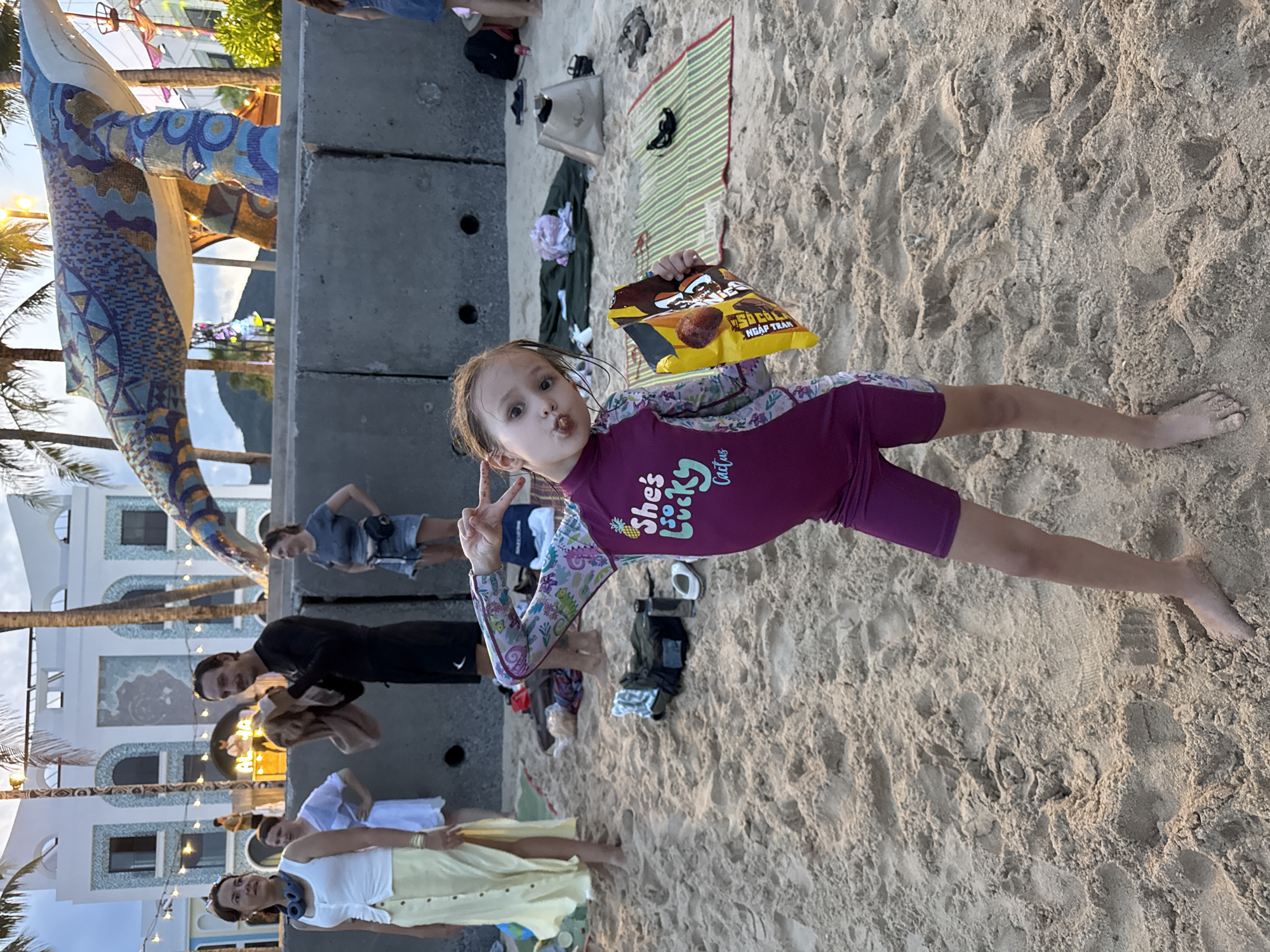 A young girl in a purple swimsuit with patterned sleeves, holding a bag of Sò ốc Nướng Tràm chips, is the focal point of a photograph taken in Bac Nha Trang Ward, Vietnam. She is lying on a sandy beach, extending one arm in a peace sign gesture. Other people are in the background. One is standing near a concrete wall, while others are further away, closer to buildings with lights and a decorative mosaic structure. There is a green mat with a black stripe on the sand nearby. The overall scene suggests a casual, outdoor gathering at or near the beach.