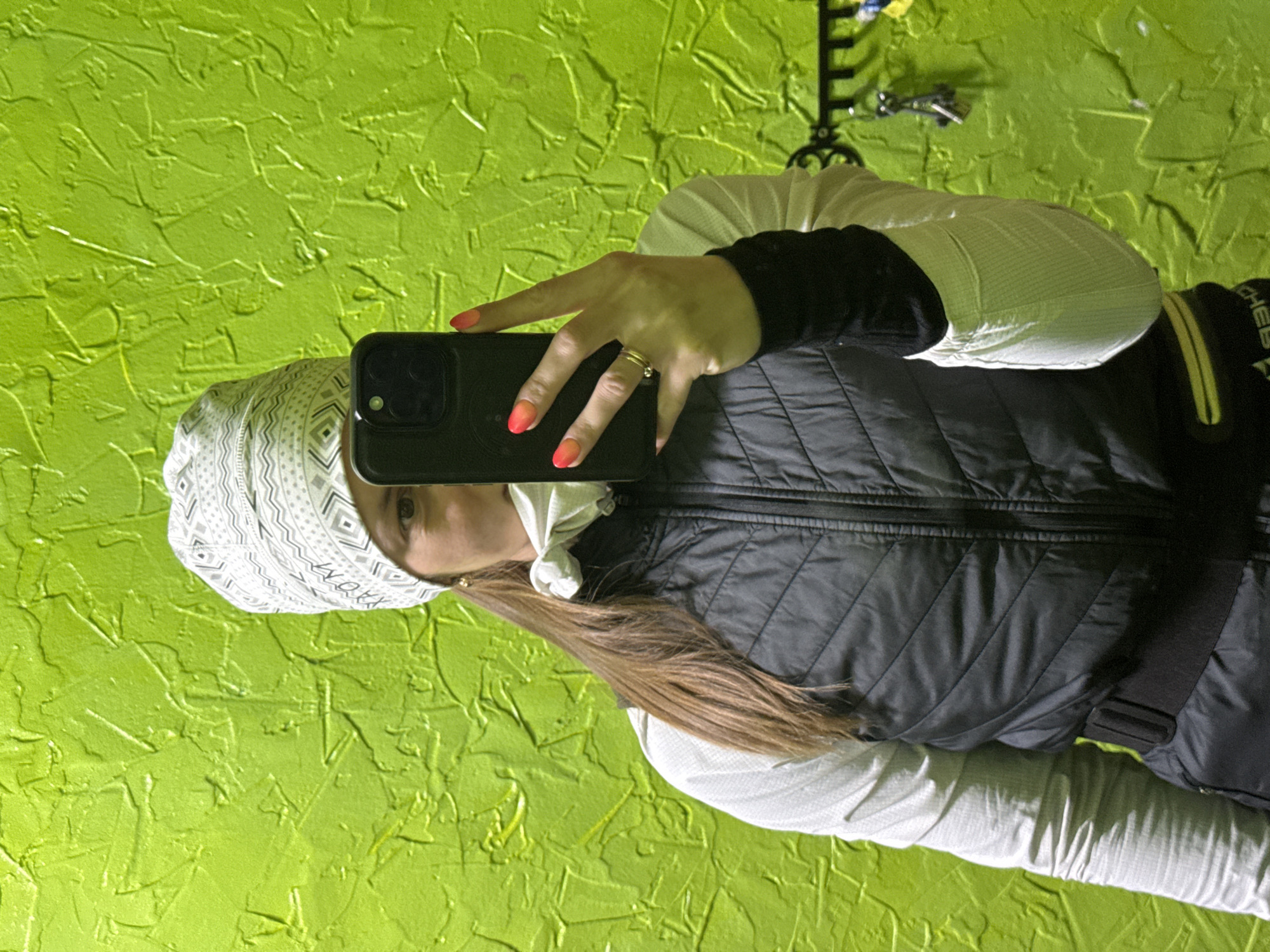 A woman is captured taking a selfie in an indoor setting, likely in Chaikovsky, Russia. She holds a dark-colored smartphone, consistent with an iPhone, in her right hand, positioned as if capturing her reflection or a self-portrait. Her nails are painted with a noticeable orange and red ombre effect, and she wears a delicate gold ring on her ring finger.
She has long, brown hair and is dressed in practical cold-weather clothing: a white long-sleeved top, a black quilted puffer vest, and a white beanie or headwrap adorned with a geometric pattern. Part of her left eye is visible, looking towards the camera.
The background consists of a bright, textured lime green wall, possibly with a stucco finish, suggesting a decorated interior. On a black strap with yellow trim on her vest, partial text is visible, clearly showing "CHES". The lighting appears to be consistent with an illuminated indoor environment.