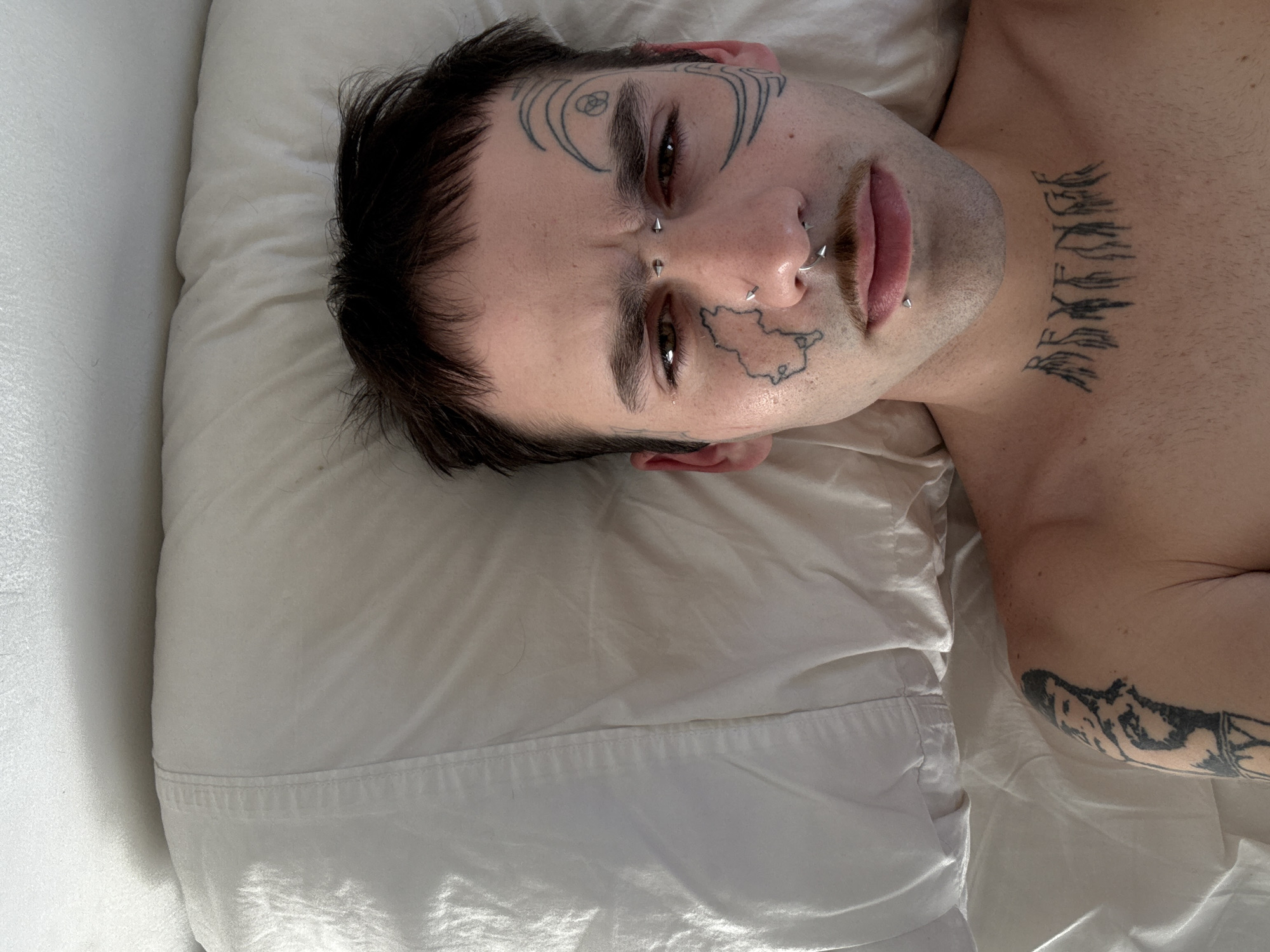 A person with dark hair is depicted lying on their back, head resting on a white pillow, looking directly at the camera. The individual has extensive tattoos and facial piercings.
On their face, abstract line patterns tattoo the skin around their eyes, a small geometric design is visible on their forehead, and the outline of a map-like shape is tattooed on their left cheek. Piercings include two spike-shaped studs on the bridge of the nose, multiple silver hoops in both nostrils, and a small stud beneath the lower lip.
A prominent tattoo on the front of the neck/collarbone area spells "REVENGE" in a stylized, fragmented font. A large, black and white tattoo, possibly depicting mountains or a landscape, is partially visible on their right arm.
The person's expression is neutral and direct, with slightly parted lips. The setting appears to be indoors, likely a bed, with white sheets and pillow illuminated by soft, natural daylight. The image is set in Buenos Aires, Argentina.