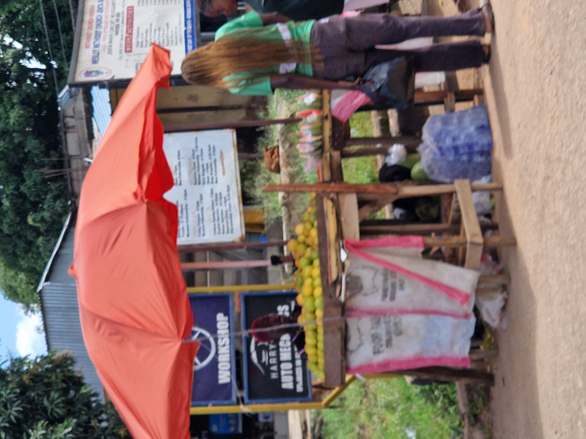 The image depicts an outdoor scene in Jos, Nigeria, likely a street market. A prominent orange umbrella shades a fruit stall laden with a large quantity of yellow citrus fruits, possibly oranges. Behind the fruit stand, a person with long, reddish-brown hair wearing a green t-shirt and dark pants is visible, carrying a black bag. Another person, partially visible, stands to the right. Signs are displayed, including one that reads "WORKSHOP" and another advertising "HARRY AUTO MECH". The environment appears to be a paved area with a grassy background, and some corrugated metal structures are visible in the distance. The overall impression is that of a bustling market setting.