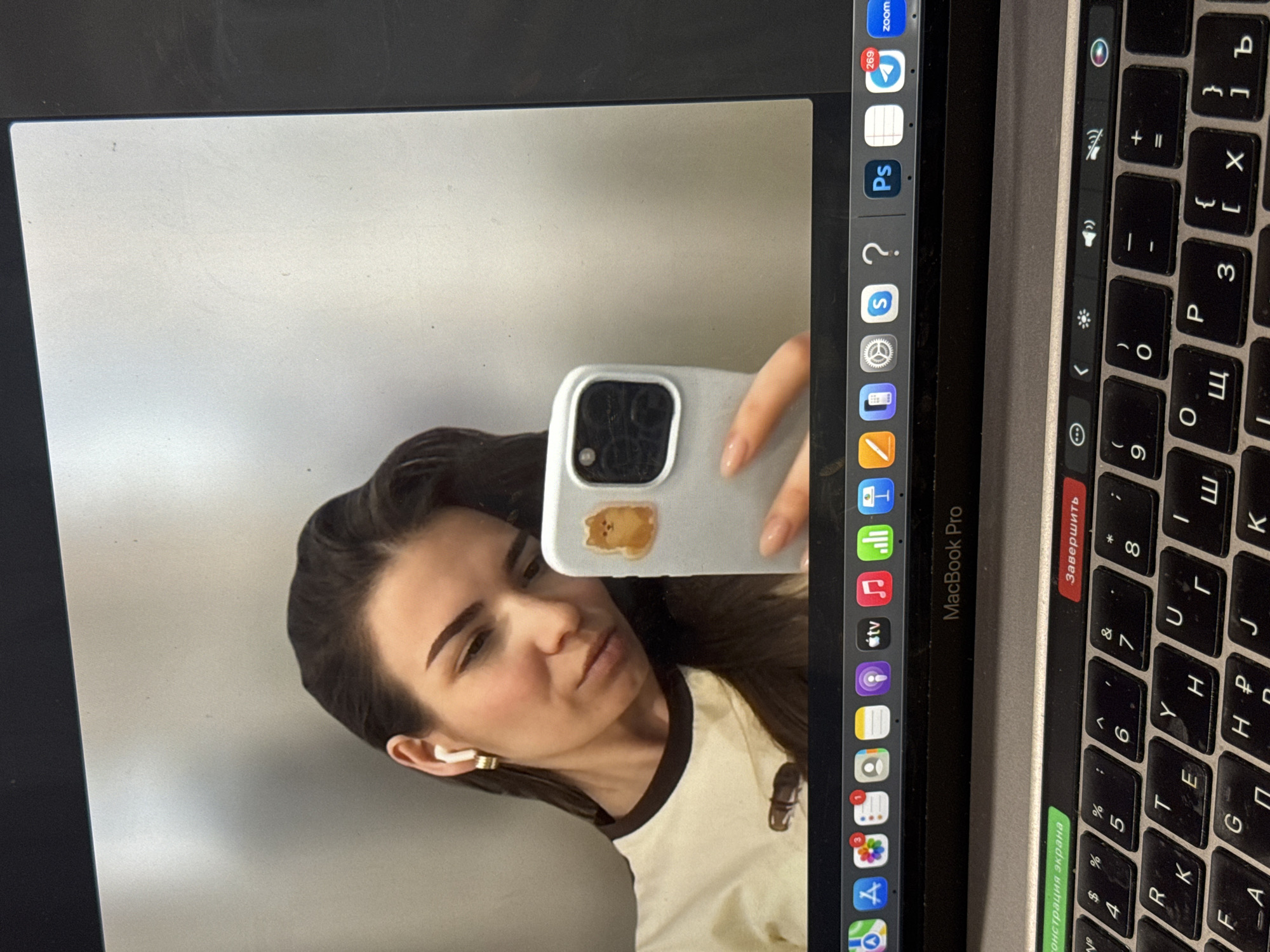 The image shows a MacBook Pro displaying a selfie of a woman. The laptop's screen is the primary focus. The woman has brown hair, is wearing a white shirt with brown trim, and has an earbud in her left ear. She is holding a white smartphone with a sticker on it. The MacBook's dock and keyboard are visible on the right side of the image, along with the top of the screen. The keyboard includes some keys labeled with Cyrillic characters. The environment appears to be indoors. The city of Khimki, Russia, is not identifiable.