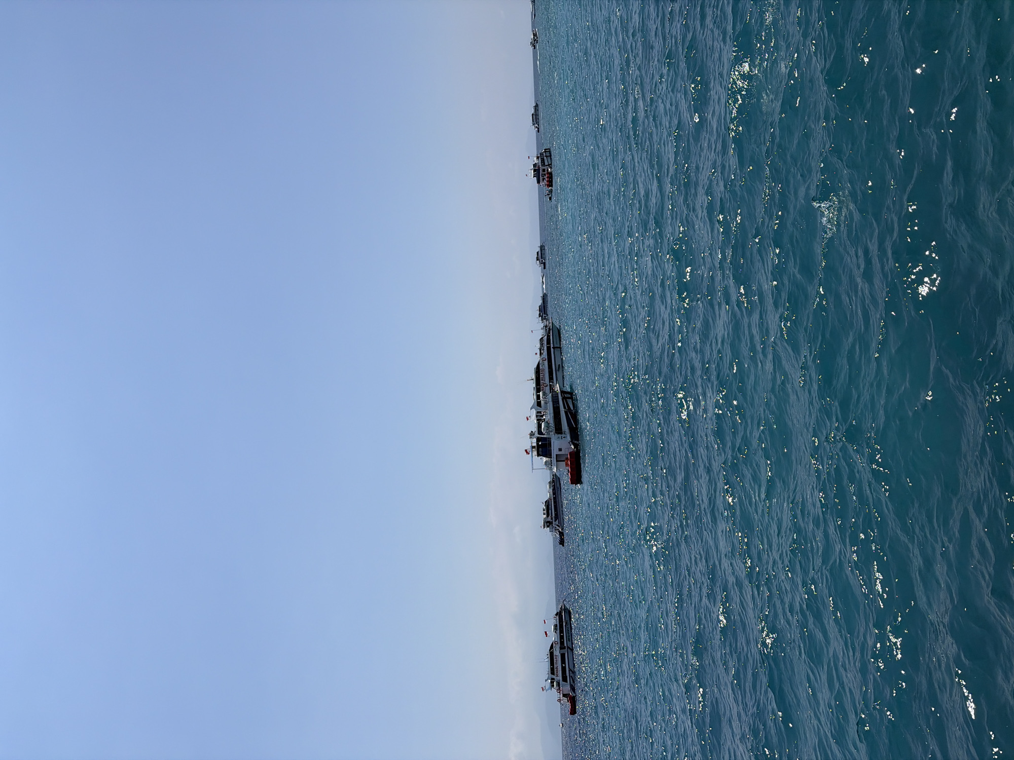 A wide shot captures a clear blue sky over a vast expanse of deep blue ocean, dotted with numerous white passenger boats. The boats, varying in size and design, are spread across the water, some appearing to be docked or anchored, while others are in motion, leaving faint wakes. Many of the boats display red and white flags. In the distance, a hazy landmass with rolling hills is visible, suggesting the presence of an island. The water is rippled, with sunlight glinting off its surface, indicating a bright, sunny day. The scene is tranquil and suggests a popular nautical area, potentially a ferry terminal or a popular spot for boat tours in Kampung Toyapakeh, Indonesia.