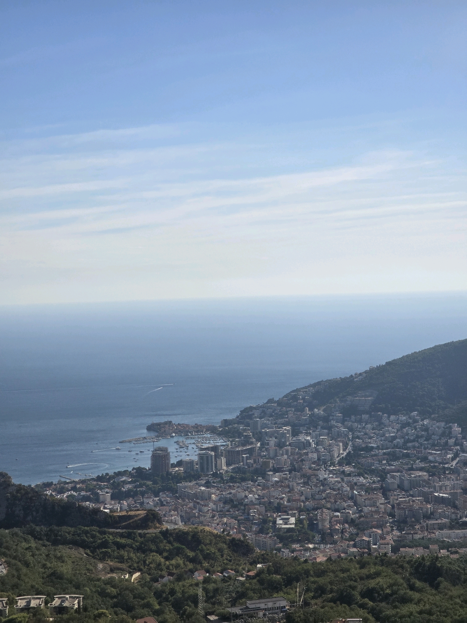 This is an aerial, wide-angle shot of the coastal city of Budva, Montenegro, as seen from a high vantage point in the hills surrounding Lapčići. The scene is dominated by the blue expanse of the Adriatic Sea to the left, meeting a hazy horizon under a pale blue sky with wispy white clouds. The foreground is covered in dense, dark green vegetation.
Descending from the hills, the cityscape of Budva unfolds. Buildings, varying in height and architectural style, are tightly packed together, their rooftops a mix of red tiles and concrete. Several modern, taller buildings, including a marina with numerous yachts and smaller boats, are visible closer to the waterfront. A prominent road snakes through the urban area, and a breakwater or pier extends into the sea. The city extends along the coastline and climbs up the adjacent hills. In the distance, on the sea, there are a few white wakes trailing small boats. The overall impression is one of a densely populated coastal town nestled between lush green hills and the expansive sea.