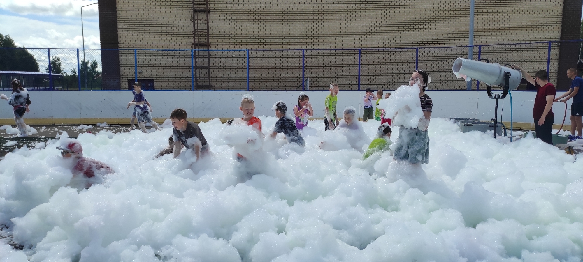 The image depicts an outdoor foam party taking place in Noviy, Russia. A large quantity of white foam fills the foreground and midground, with numerous children playing and interacting within it. Some children are running, while others are sitting or lying down in the foam. They are dressed in casual clothing, and many appear to be enjoying themselves. In the background, there is a building with a brick facade and a fence. A large foam machine is visible on the right side of the image, operated by two adults. The setting appears to be an open area, possibly a park or a recreational facility. The sky is partly cloudy, indicating daytime.