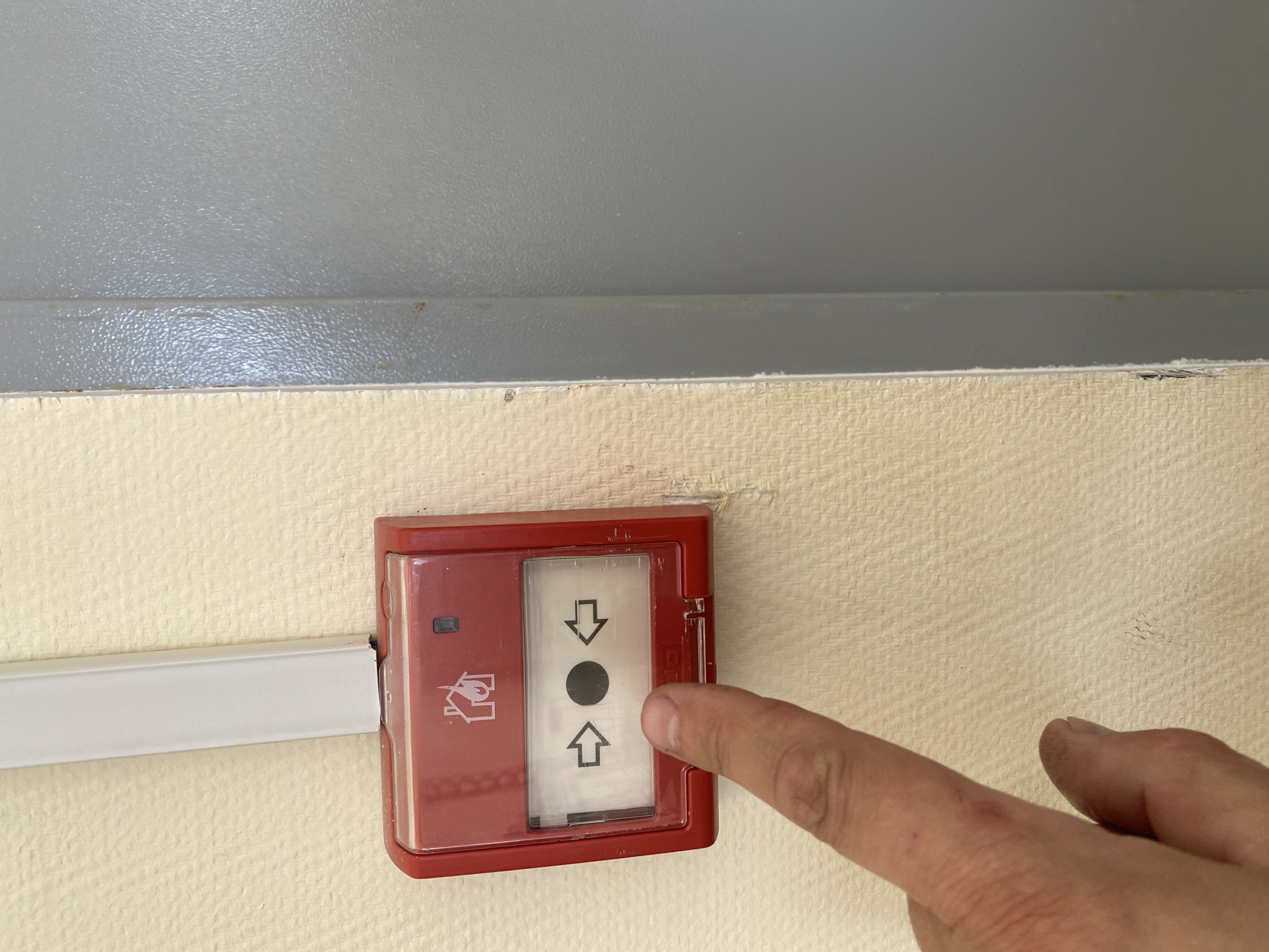 A close-up, eye-level shot shows a human hand interacting with a red fire alarm call point mounted on a light-colored, textured wall. An index finger is positioned over the clear plastic cover of the alarm, appearing to press or prepare to press it. The rectangular red casing of the fire alarm features a transparent panel with a black circular element in its center, flanked by an upward-pointing arrow below and a downward-pointing arrow above, indicating an activation mechanism. To the left of this clear panel, on the red casing, a small black square (possibly an indicator light) and a pictogram depicting a house with flames (a common fire alarm symbol) are visible. A white cable trunking or conduit runs horizontally along the light-colored wall, leading into the left side of the fire alarm unit. Above the light-colored wall, a grey wall with a similar textured surface is present, separated by a horizontal line or molding. The setting appears to be indoors, possibly in a public or commercial building. No specific time of day is discernible, and no text is clearly legible on the device, only symbols.