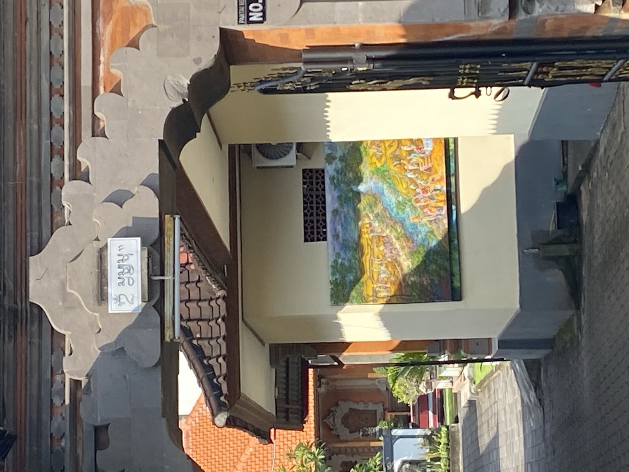 A view from above, likely an upper floor of a building, looks through a narrow passage between two buildings in Nusa Dua, Indonesia. The building to the left appears older, with intricate stonework. There is a sign with text in a local language. A tiled roof with red tiles and brown eaves is visible. The building to the right has a large, colorful painting on the wall. There is an air conditioning unit mounted on the wall above a decorative vent. Beyond the buildings, more of the city is visible, including greenery, buildings, and a paved area. The view suggests a densely built area with traditional Balinese architecture blended with modern elements.