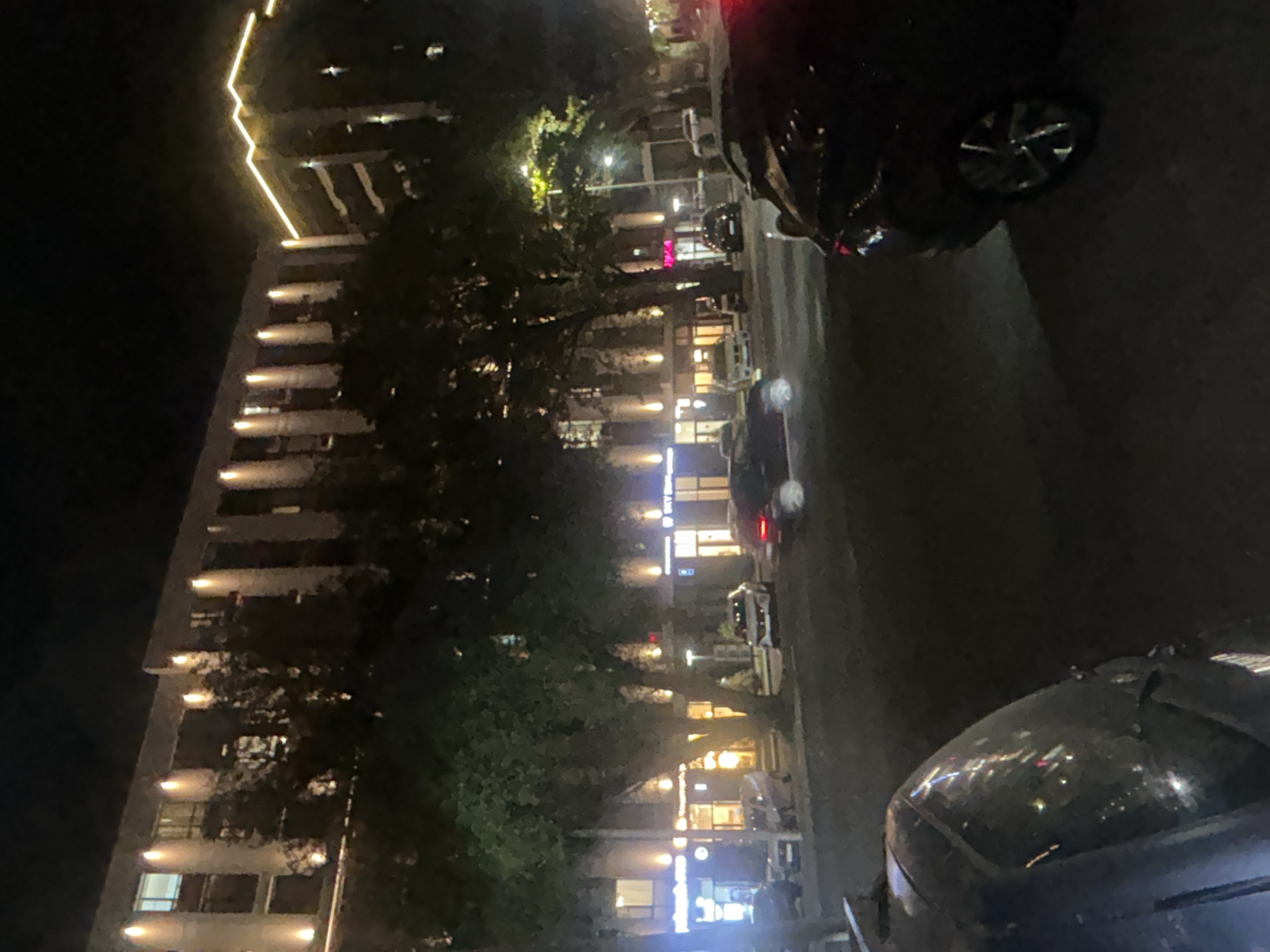 The image captures a nighttime street scene in Almaty, Kazakhstan, with a multi-story building dominating the background. The building features numerous windows and is illuminated by exterior lights and a strip of warm-toned lights along its upper edge. In front of the building, several trees with dense foliage are visible, their branches extending towards the street. The street itself is lined with parked cars and a few vehicles in motion, their headlights and taillights casting reflections on the wet or polished surface of the road. The foreground is occupied by the rear end of a dark-colored car, its wheel and part of the body clearly visible. Several storefronts with illuminated signs are discernible along the lower level of the building, suggesting a commercial area. The overall atmosphere is dark, with artificial lights from the building and vehicles creating pockets of illumination.