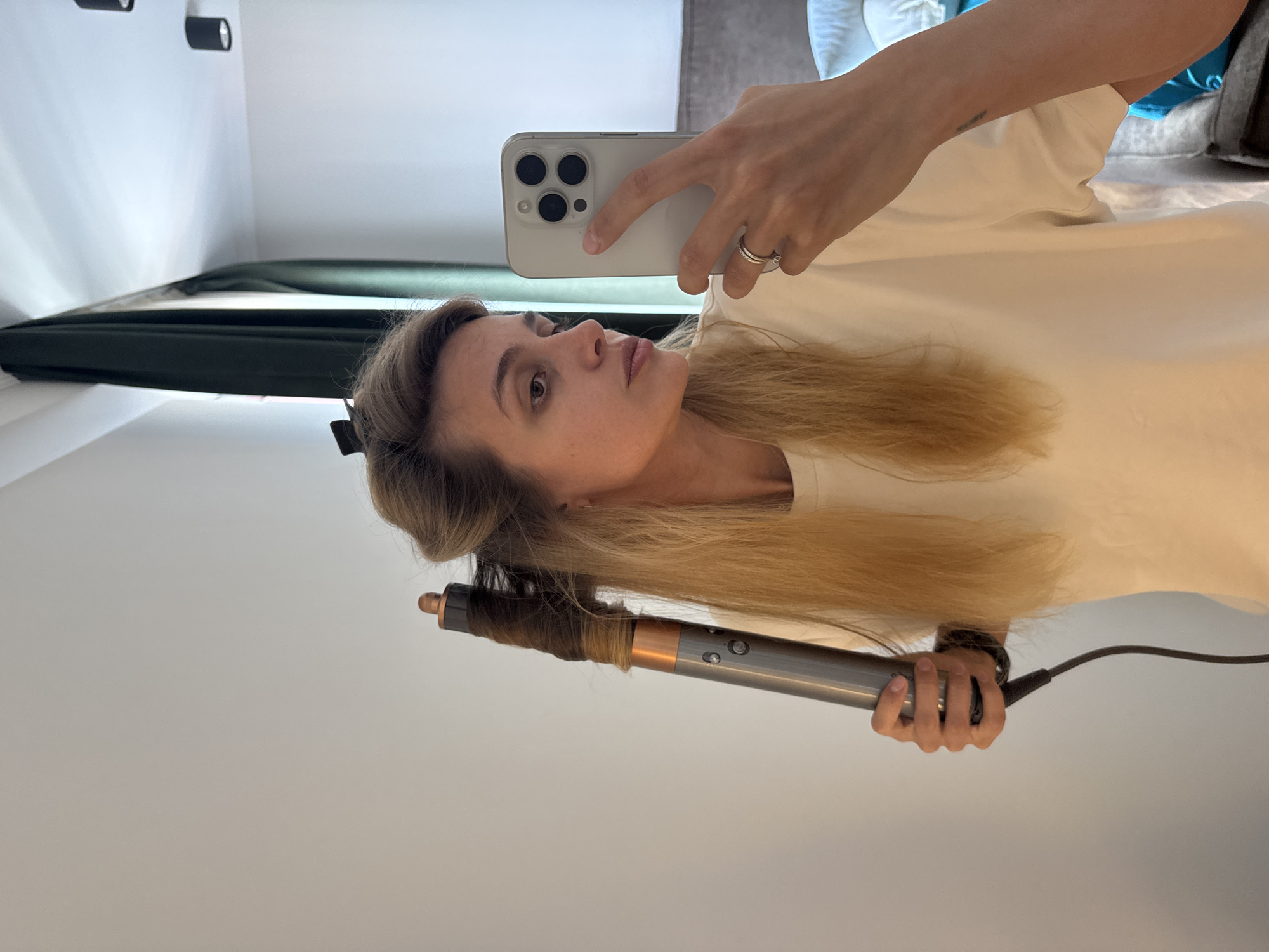A person is holding a cell phone with multiple camera lenses in their right hand, taking a selfie while using a Dyson Airwrap on a strand of their light brown hair. The person is wearing a light-colored, possibly white, t-shirt. The background includes a white wall and a dark-colored curtain or window covering. The location appears to be indoors.