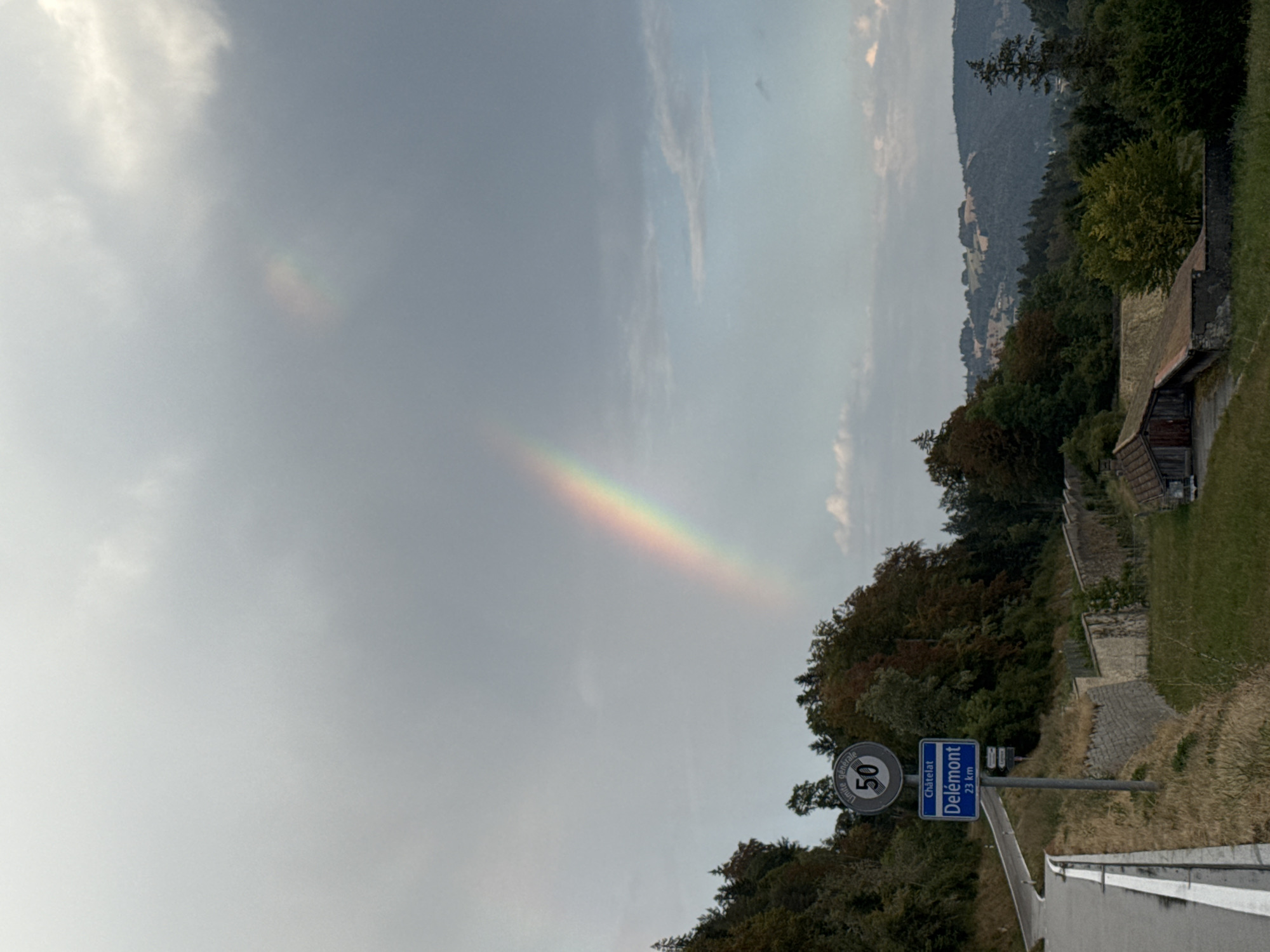 The image shows a road curving up a hillside, with a sign indicating "Châtelat / Delémont 23 km". A speed limit sign of 50 is also visible. The sky is cloudy with a rainbow visible. Trees line the hillside, with some autumnal colors. In the foreground, there is a stone wall and a small building. In the background, there are rolling hills and more trees. The scene appears to be a rural landscape in the vicinity of Bellelay, Switzerland.