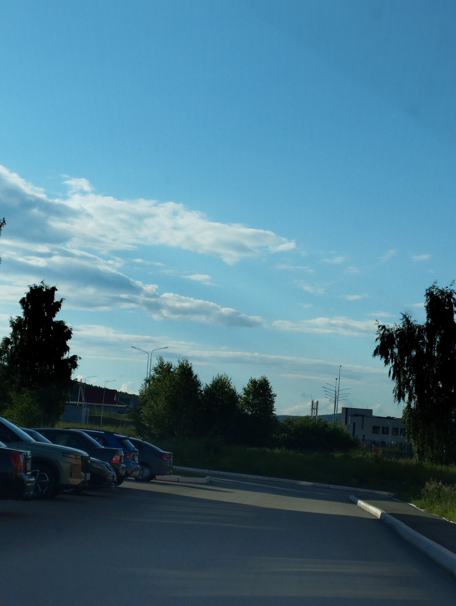 The image captures a parking lot scene under a clear blue sky with scattered clouds. Several cars are parked in a row, suggesting a daytime setting. The cars appear to be facing away from the viewer, with their rear ends visible. The foreground consists of an asphalt surface, likely a road or parking area, with a white curb. In the background, lush green trees line the edge of the parking lot, and beyond them, a building with a modern facade can be seen. The overall atmosphere is calm and ordinary, with no discernible activity or people in the frame. The lighting suggests it's daytime, possibly late afternoon, due to the angle of the shadows and the warm tones in the sky. The weather appears pleasant, with clear skies and some clouds adding visual interest. No text is visible in the image.
