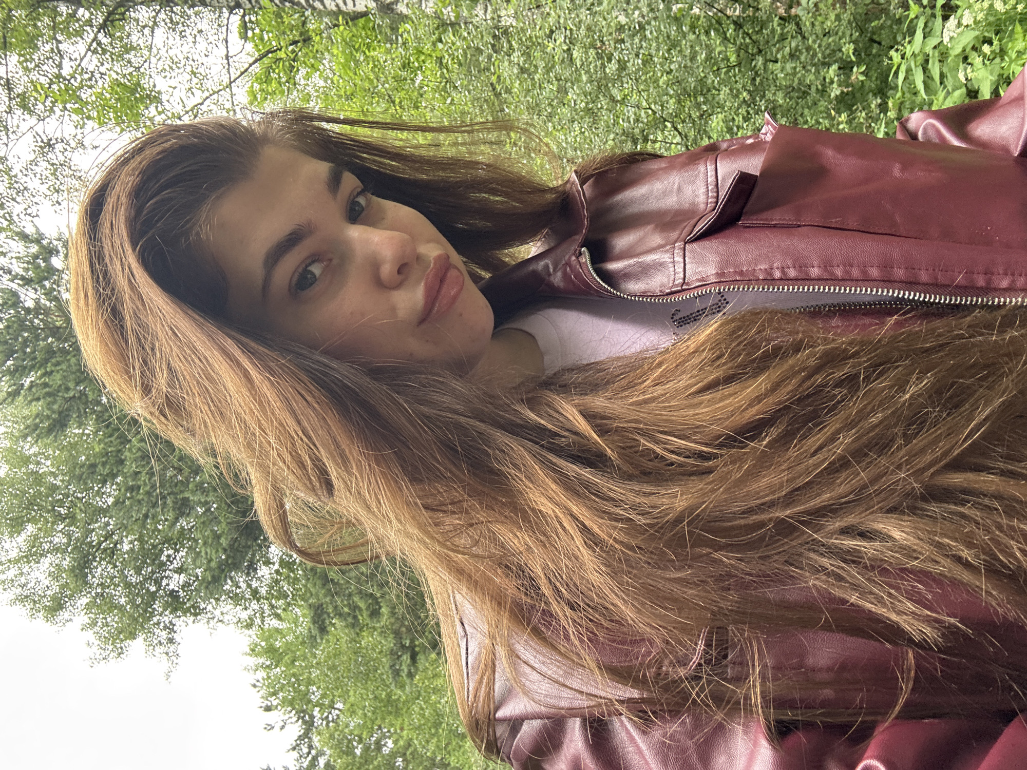 A young woman with long, wavy brown hair is taking a selfie outdoors. She is wearing a maroon leather jacket over a white t-shirt. Her hair is covering the left side of her face and shoulder. She has light-colored eyes and a neutral expression. The background consists of lush green trees and foliage, suggesting a natural or park-like setting. The lighting appears to be overcast, with diffused daylight. The specific location is not discernible from the visual information provided, but the prompt mentions Мещёры, Russia, which is a region known for its forests.