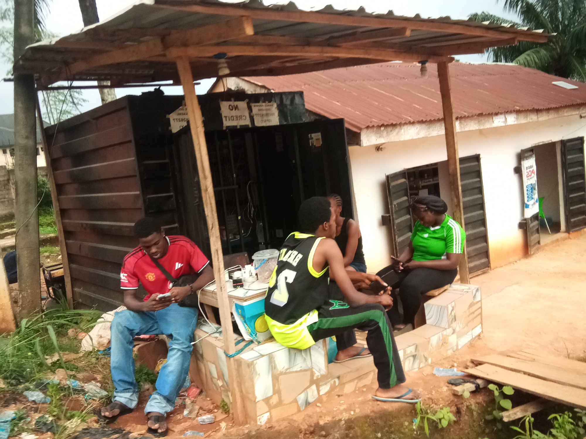 In Oko Town, Nigeria, a low-slung, open-air structure with a corrugated metal roof supported by rough-hewn wooden beams stands in front of a white building with dark-shuttered windows. The structure appears to be a makeshift shop or stall. Several people are present. A young man in a red Manchester United jersey and blue jeans is seated on a low concrete platform, looking at his phone. To his right, another young man in a black and neon yellow basketball jersey and black and green pants is also seated on the platform, interacting with a smartphone. A woman in a green and white striped shirt and black pants is seated next to him, also looking at a phone. A fourth person, whose face is partially obscured, is seated further to the right. The ground around the structure is dirt and covered in debris, including plastic bags and discarded items. The environment suggests a humid, possibly tropical setting with lush green foliage visible in the background and a palm tree to the left. A small wooden bridge crosses a ditch or drainage channel in the foreground. Signs on the structure include "NO CREDIT" in English, suggesting it is a commercial establishment.