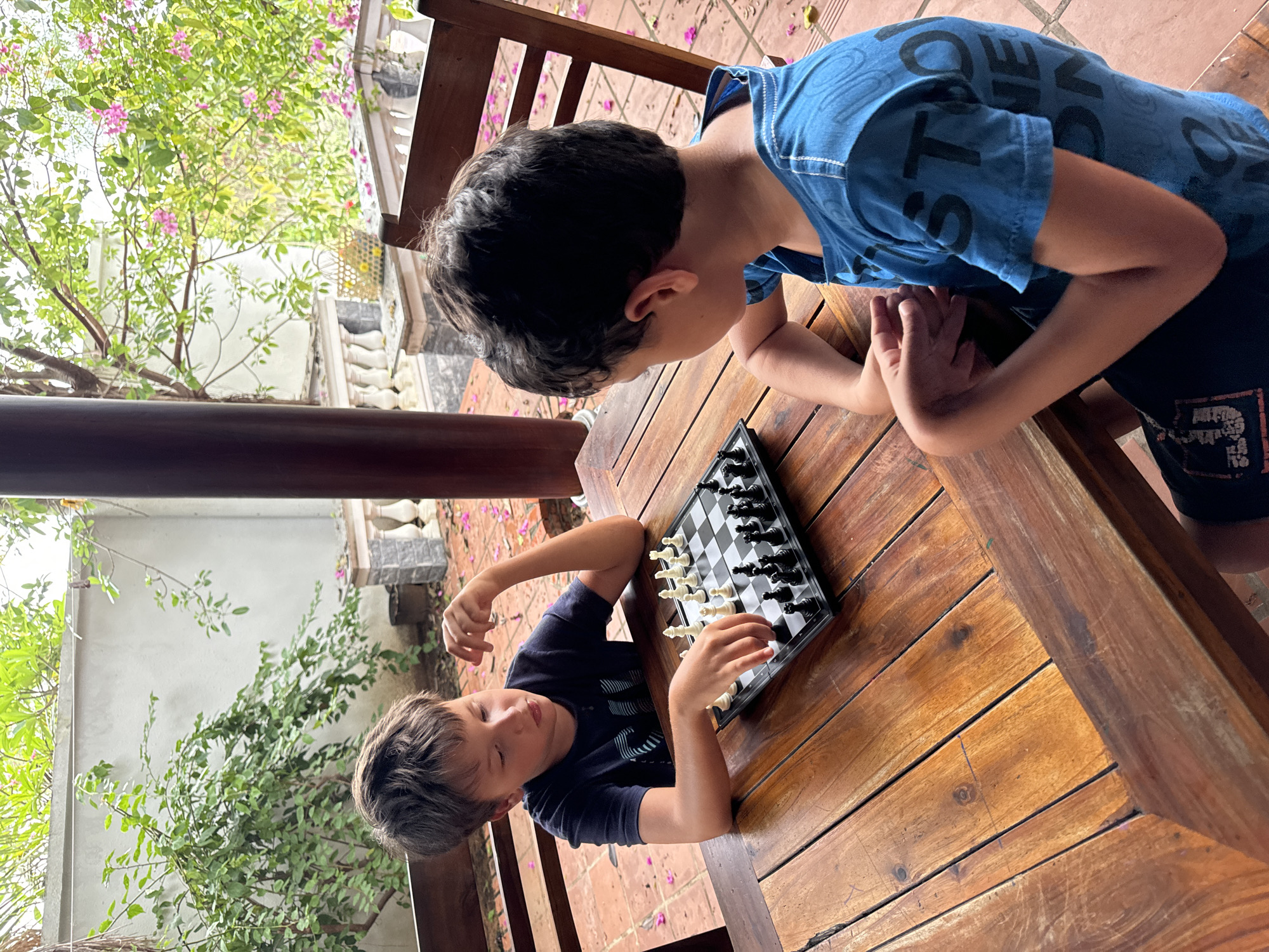 Two boys are playing chess on a wooden table outdoors. The chessboard is set up with pieces. One boy wears a blue patterned t-shirt, the other a darker blue patterned t-shirt. The scene appears to be in a garden or courtyard, possibly in Mũi Né, Vietnam, given the presence of tropical vegetation. The backdrop includes foliage, trees, and part of a building with a white facade. The table and chairs are made of wood.