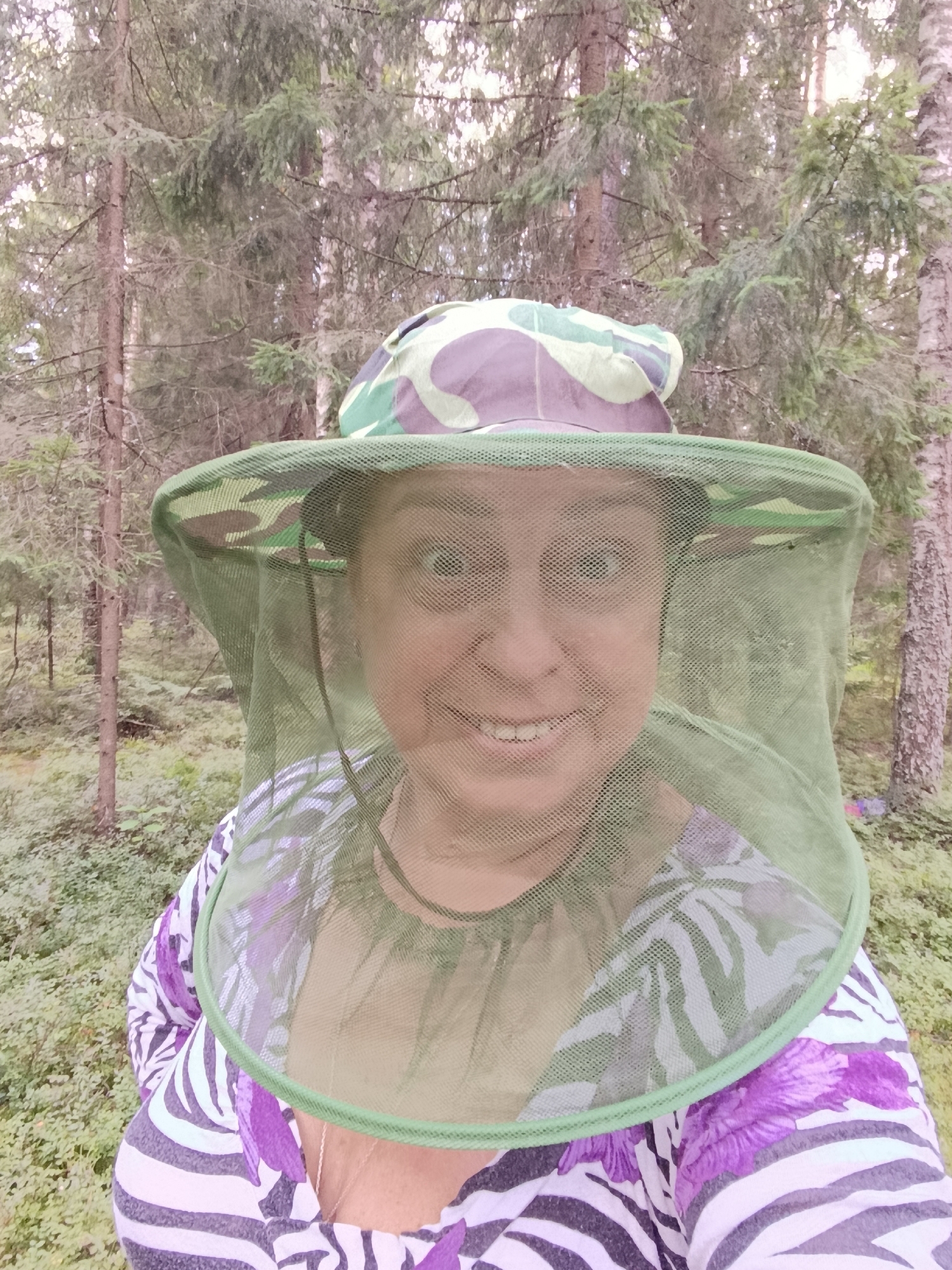 A woman with a light complexion and dark brown hair, wearing a camouflage-patterned hat with a green net that covers her face and neck, is standing in a forest. She is smiling, showing her teeth. She is wearing a purple and white zebra-striped shirt with a floral pattern overlay. The trees in the background are tall and slender, with sparse foliage, indicating a temperate or boreal forest environment. The ground is covered in low-lying green vegetation. The lighting is soft and diffused, suggesting it is daytime. The scene is a selfie taken by the woman herself.