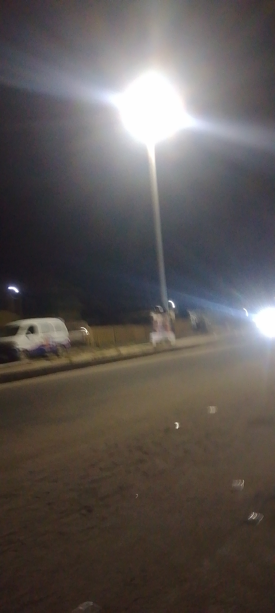 The image depicts a nighttime outdoor scene on a city street in Unknown, Unknown. An intensely bright street light, centrally positioned and overhead, illuminates the immediate area and causes significant lens flare. The sky is dark.
The foreground and midground show an asphalt road surface with faint reflections and scattered small, light-colored specks, possibly litter. A curb or raised divider runs parallel to the left side of the road. Beyond this, a light-colored panel van, possibly white with indistinct blue or purple markings on its lower body, is visible. Further along the left side, behind the van, is a dark fence or barrier. To the right of the van, on what appears to be a sidewalk or shoulder, a large, rectangular object, possibly a banner or signboard, stands upright. Its surface is light-colored with dark, indistinct shapes. In the distance, multiple bright light sources, consistent with vehicle headlights or streetlights, are present. No individuals are discernible. The overall image is blurry due to low light conditions or camera motion.