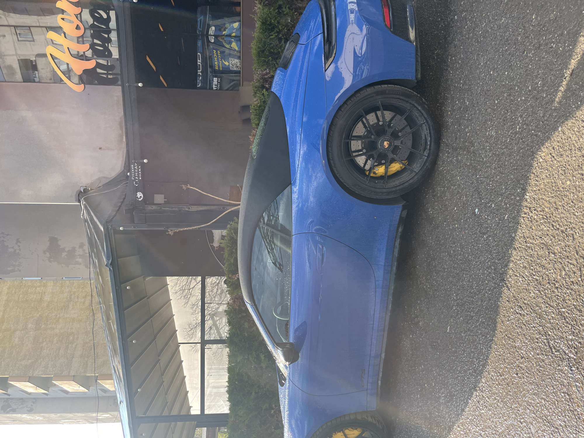 A blue Porsche sports car is parked on a paved surface, possibly a street or parking lot, next to a building. The car's black rims and yellow brake calipers are visible. The building has a sign in a Cyrillic alphabet and includes the word "vodka". Trees are partially visible behind the building. Based on these signs, the location appears to be Sofia, Bulgaria. The car seems to have been recently washed, with water droplets on its surface. The scene is photographed in daylight with strong sunlight creating shadows.