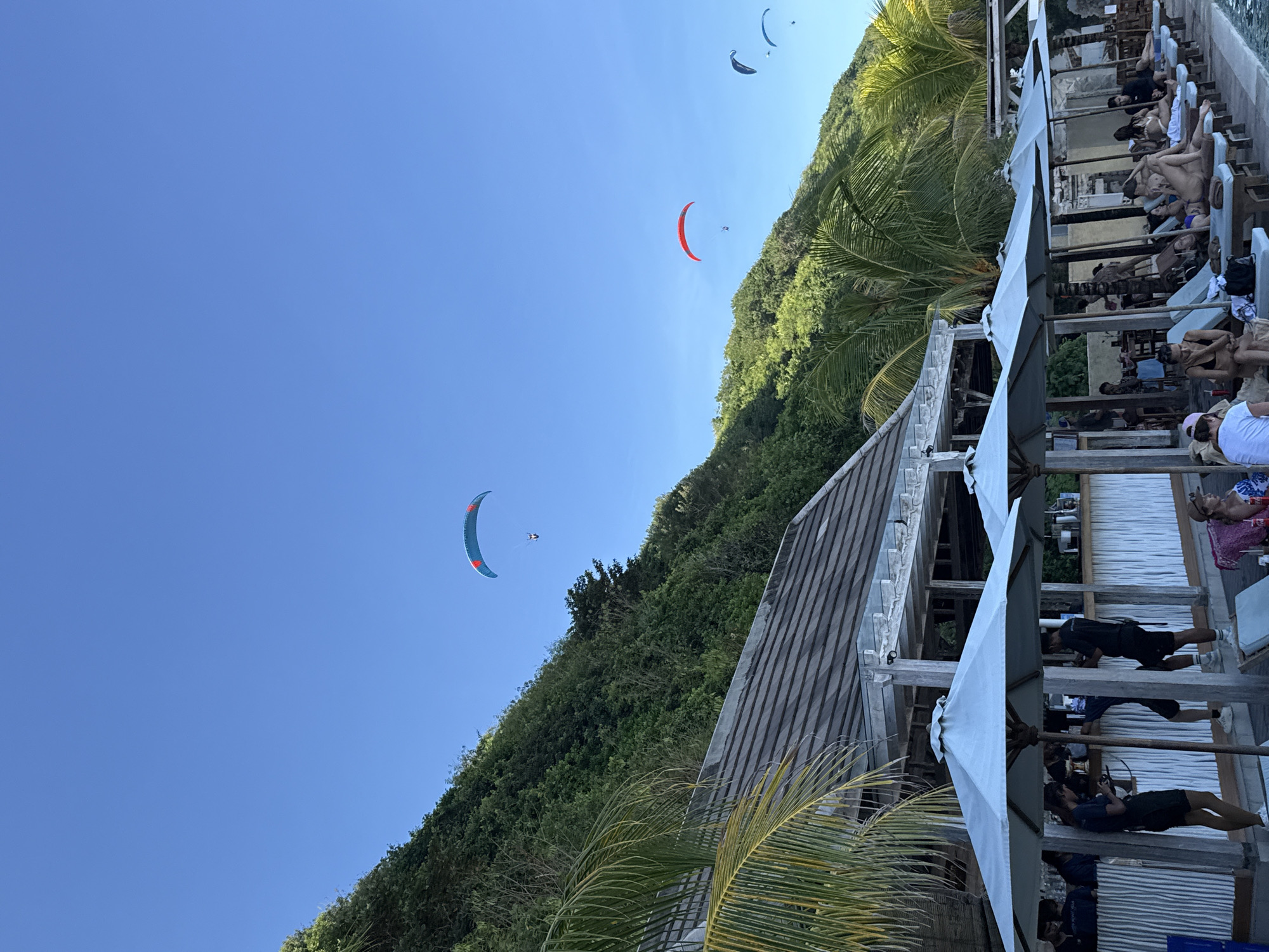 The image, taken in Kuta Selatan, Indonesia, shows a bright, clear, blue sky with three paragliders in the air, two visible and one faintly in the upper right corner. Lush green foliage covers a steep hillside that extends along the right side of the image. A wooden structure with a grey roof and white supports sits below the hillside, partially obscured by palm fronds. Adjacent to the wooden structure, a row of lounge chairs with blue cushions, shaded by white umbrellas, is occupied by people relaxing and sunbathing. Some people are partially visible in the background, behind the lounge chairs.