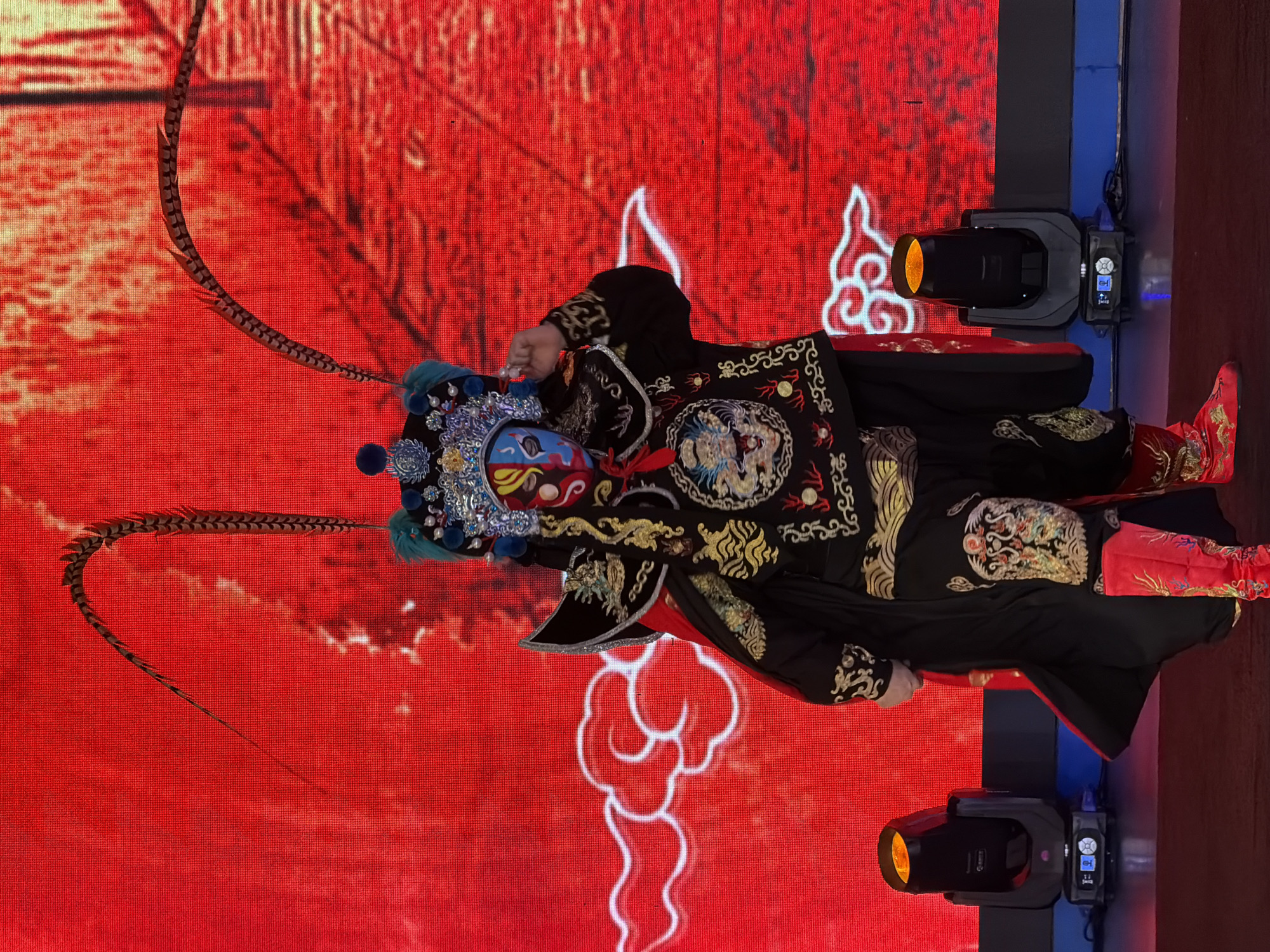 An individual in elaborate Peking Opera attire, including a mask with blue and red painted features, a headdress with long feathers, and a black and gold embroidered costume, stands on a stage. The backdrop is a vibrant red screen displaying a stylized white cloud pattern. Two stage lights, one on each side of the performer, are visible, emitting yellow light. The scene appears to be in Yuzhong District, China.