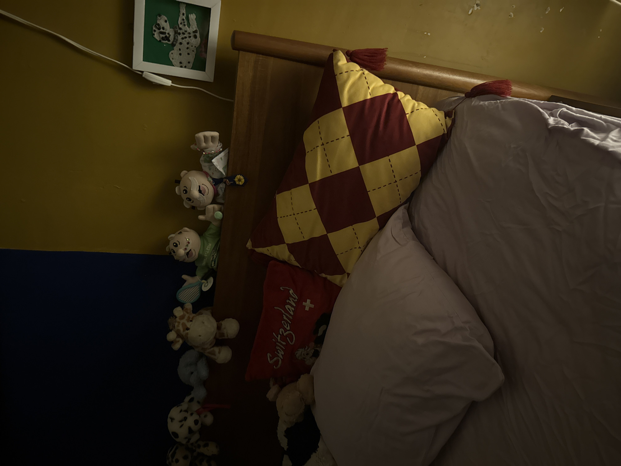The image depicts a bedroom scene. A bed with a wooden headboard is positioned with pillows and a duvet. A yellow and maroon checkered pillow with a tassel rests against the headboard. A red pillow with "Switzerland" embroidered on it is also visible. Stuffed animals are arranged along the left side of the bed, adjacent to a two-tone wall (yellow on top, blue on the bottom). A framed picture hangs on the yellow section of the wall. The scene appears to be indoors with limited lighting. There are no people present in the image. The location in Xochitepec, Mexico is not discernible from the image.