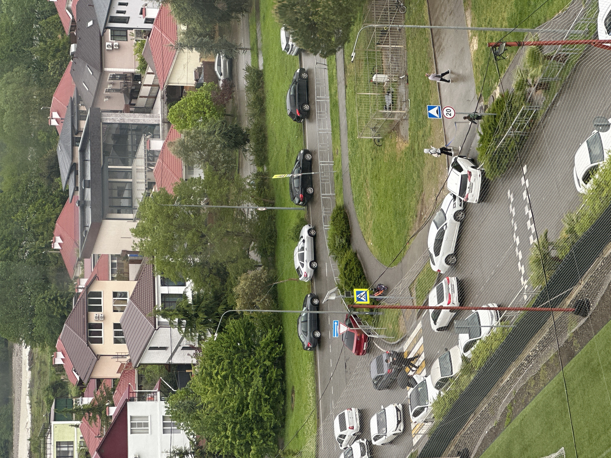 An overhead shot depicts a streetscape in Sochi, Russia, featuring a road lined with parked cars, interspersed with grass and pedestrian walkways. In the foreground, a chain-link fence obscures part of the view. Vehicles of various colors—white, black, and red—are parked along the road. Pedestrians are present, with a few individuals near a marked crosswalk, recognizable by its yellow and blue signage. Houses with red and dark-colored roofs border the street, accompanied by foliage. Street signs are visible along the road, and the setting suggests a residential area.