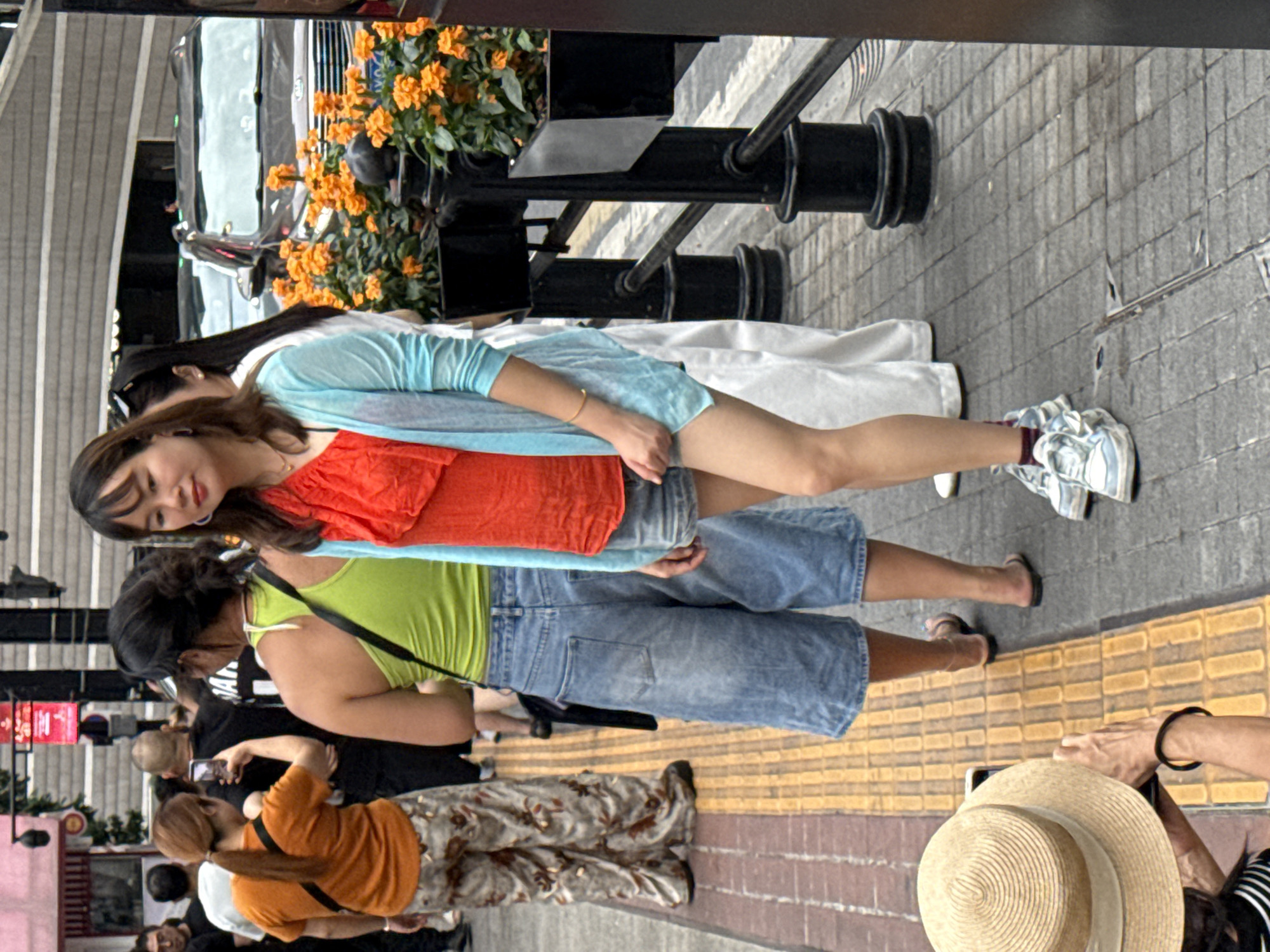 In Yuzhong District, China, a group of individuals are walking on a sidewalk next to a street with parked cars and flower planters. A woman in the foreground is wearing a red top and a light blue cardigan over denim shorts and white sneakers. To her left, another woman in a bright green tank top and denim shorts is facing away from the camera, holding a black bag. Further to the left, a woman in an orange t-shirt and patterned pants is taking a photo with her phone. Behind them, other people are visible, some walking and others standing. The environment suggests an urban setting with a mix of commercial and pedestrian activity.