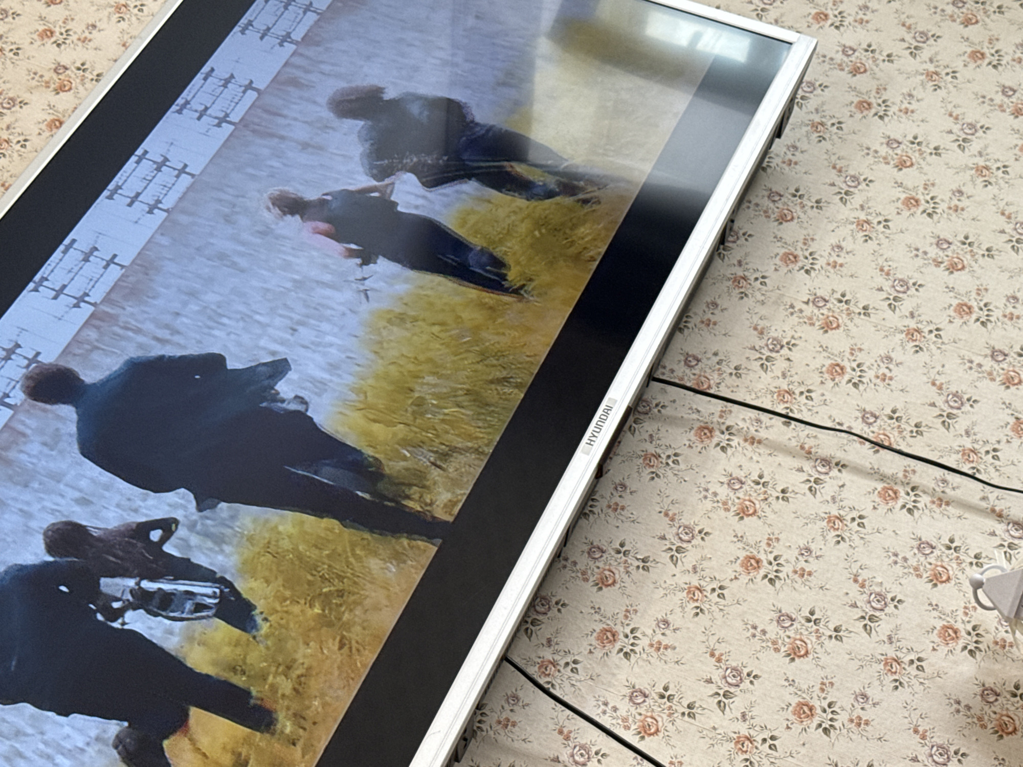 A television screen shows a scene in a likely historical setting with several figures in dark clothing. One figure is aiming a weapon, potentially a gun, towards others. The environment features elements of grass or fields and what could be a structure like a bridge in the background. The television is on a surface covered with a floral patterned cloth. The TV itself is a Hyundai brand. The location is not discernible from this image.