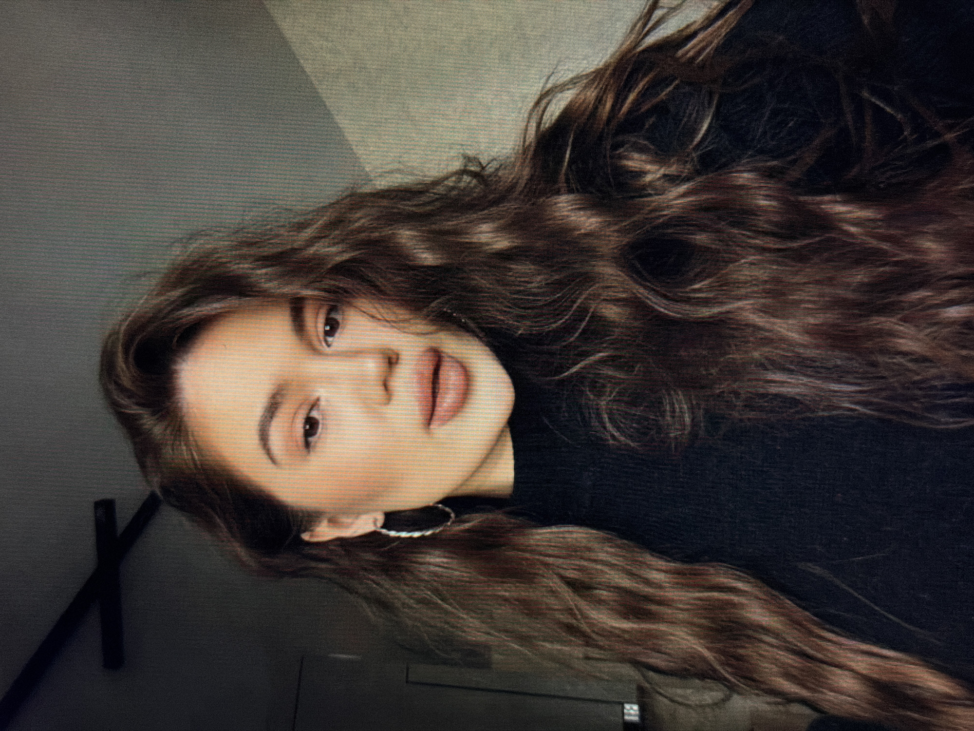 A close-up, rotated image shows a young woman with long, wavy brown hair. She is wearing a black top and a hoop earring in her visible ear. Her makeup is natural, with emphasis on her lips. The background is a textured, neutral gray wall. A dark, cross-shaped object is visible in the lower left corner of the frame, casting a shadow. The overall lighting suggests an indoor setting, possibly during the day given the diffused light. The woman's expression is neutral, with her gaze directed slightly away from the camera. No text is visible in the image. The scene appears to be a casual portrait, possibly taken in a home or studio environment. The texture of the screen on which the image is displayed is visible throughout.