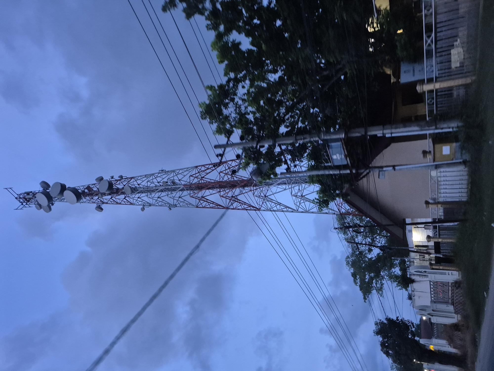 The image shows a telecommunications tower in Pantegurah, Indonesia. The tower is red and white and has several disc-shaped antennas at the top. Power lines run from the tower to the nearby buildings. The sky is cloudy and it appears to be dusk or dawn. There are buildings and trees in the background. The road is at the bottom of the image, indicating the perspective is from ground level.