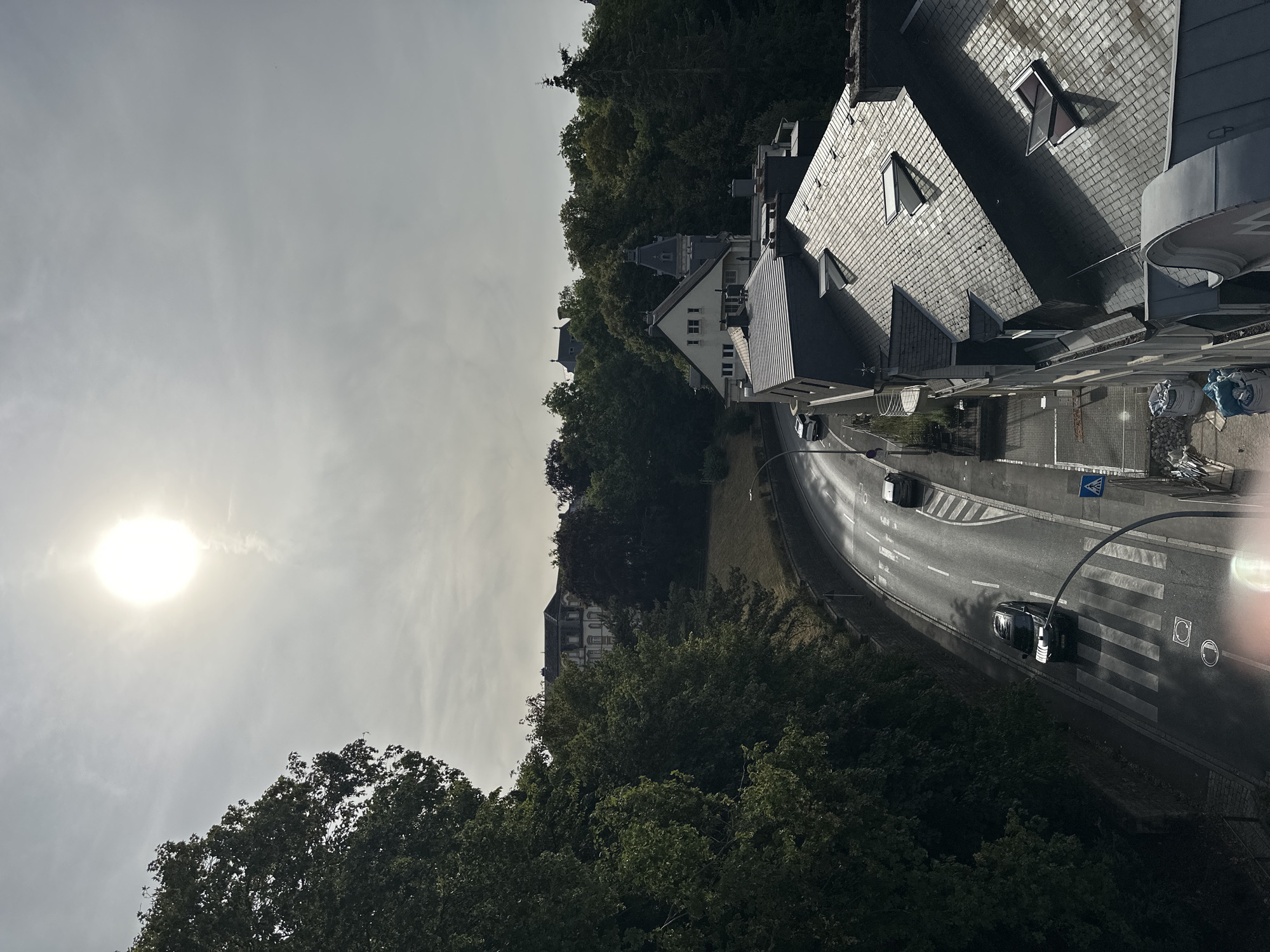 The image is an elevated, exterior, wide shot of a street scene in Luxembourg City, Luxembourg. The sun is high and bright in the hazy sky, casting a glare over the scene. Lush green trees line the left side of a curved road, which is wet from recent rain, reflecting the sunlight. Two cars are visible on the road: a black SUV driving over a pedestrian crossing and a smaller dark car further up the road. A white pedestrian crossing is clearly marked on the road. On the right side of the street, a multi-story building with a slate roof and several dormer windows is prominently featured. A streetlamp curves over the road near the pedestrian crossing. In the background, more trees and the roofs of other buildings are visible on a hillside, with a large, stately building at the top of the hill. The scene depicts a quiet street with moderate traffic and residential buildings surrounded by abundant greenery.
