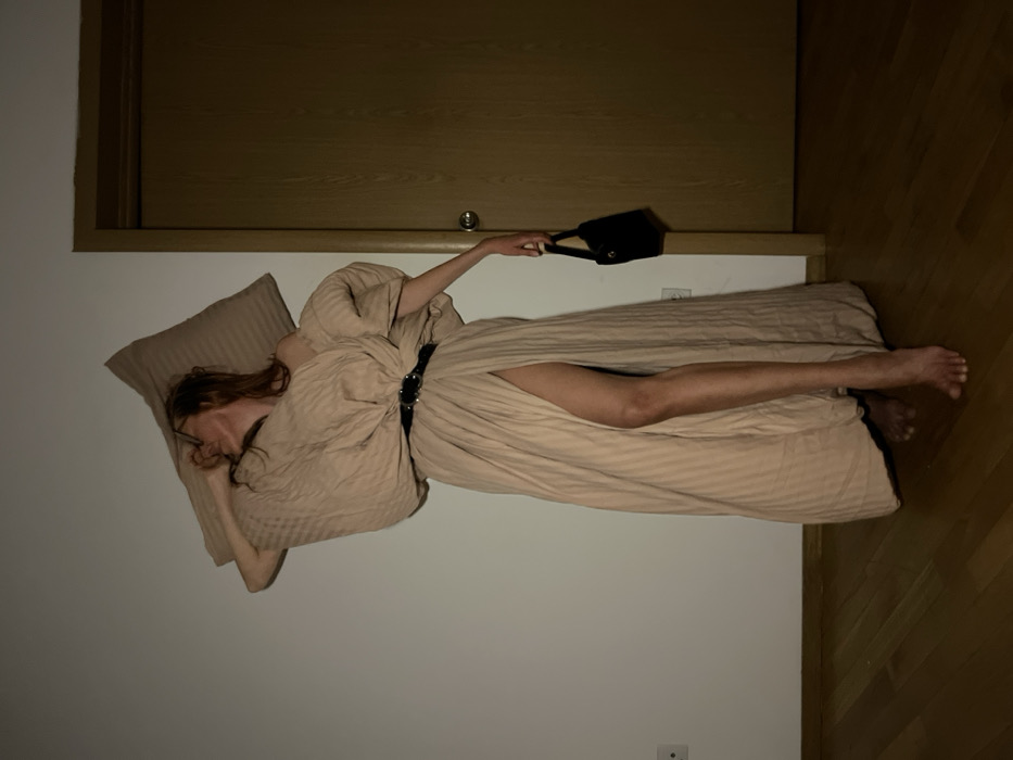The image depicts a single individual, presumably female, standing barefoot in an indoor residential setting attributed to the city of Budva, Montenegro. The setting includes a plain off-white wall to the left and rear, and a light brown wooden floor. A light brown wooden door with a metallic doorknob is visible on the right side of the frame. An electrical outlet with two circular receptacles is present on the lower left portion of the wall.
The individual possesses light brown or auburn hair and is wearing glasses. Their head is tilted back and to the right, with the mouth partially open, suggesting an exhalation or vocalization. They are dressed in a makeshift garment fashioned from a beige, subtly striped fabric, resembling a duvet cover or sheet, which forms a long, flowing dress with voluminous sleeves and a high slit on the left side, exposing the entire left leg. A black belt with two silver-toned circular buckles cinches the garment at the waist. A matching beige, striped rectangular pillow is positioned on the individual's head and right shoulder. The left arm hangs loosely, holding a small, black handbag with a short handle. The lighting appears to be artificial, casting subtle shadows.