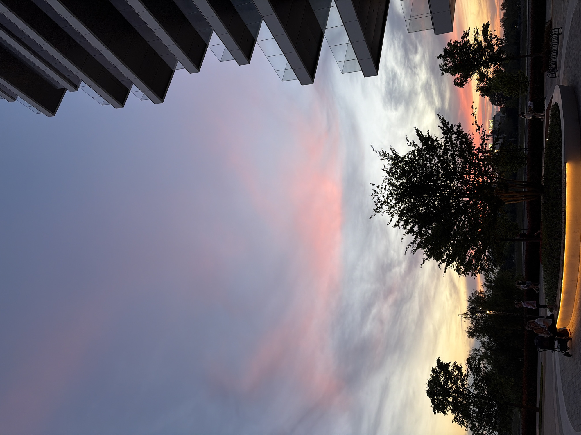 The image shows a view looking up at a sunset sky framed by a modern building on the left and trees and a walkway on the right. The sky transitions from blue to pink and orange hues, indicating sunset. The building has a layered, overhanging design with glass elements. On the right, silhouettes of trees are visible against the colorful sky, along with a lit walkway or pathway. People are indistinctly visible on the pathway, suggesting they are walking or relaxing. The location is Belgrade, Serbia.