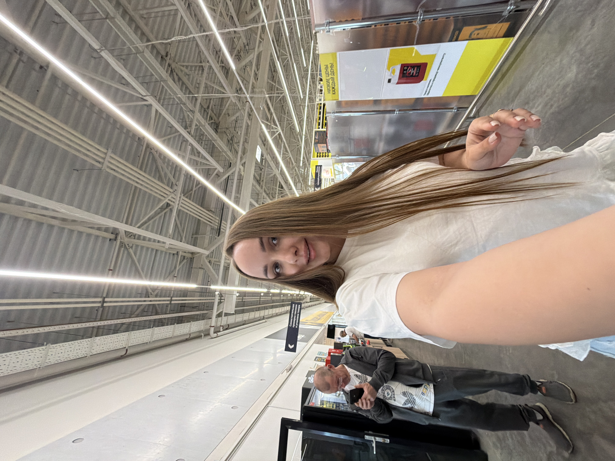A selfie photograph captured in a commercial environment, likely a large retail store or warehouse in Izhevsk, Russia. The photograph features a woman with light brown hair wearing a white t-shirt and a man, presumed to be older, in a grey suit jacket over a graphic t-shirt, along with dark pants and shoes. The man appears to be looking at or using a mobile phone. The backdrop reveals a high, industrial ceiling structure with fluorescent lighting, shelving units, and aisle signage in Russian script. The overall setting is characterized by wide aisles and commercial fixtures suggestive of a bulk retail environment.